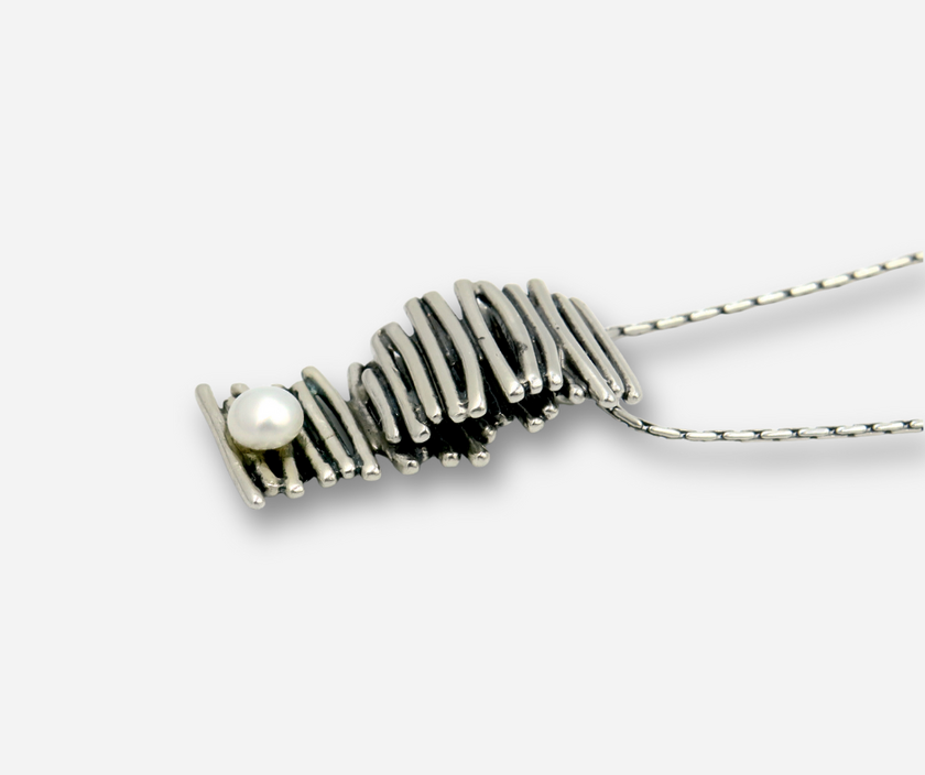 שרשרת בסגנון ארכיטקטוני עשויה כסף סטרלינג, עם תליון בעיצוב ייחודי – מעין גליל שנוצר מליפוף חוטי כסף בצורה לא סימטרית, אך הרמונית. בבסיס הסליל שובצה פנינה קטנטנה שמרככת את המראה הכללי ויוצרת מוקד אור קטן. פריט אומנותי שמתאים לאוהבות עיצוב לא שגרתי ומעודן.