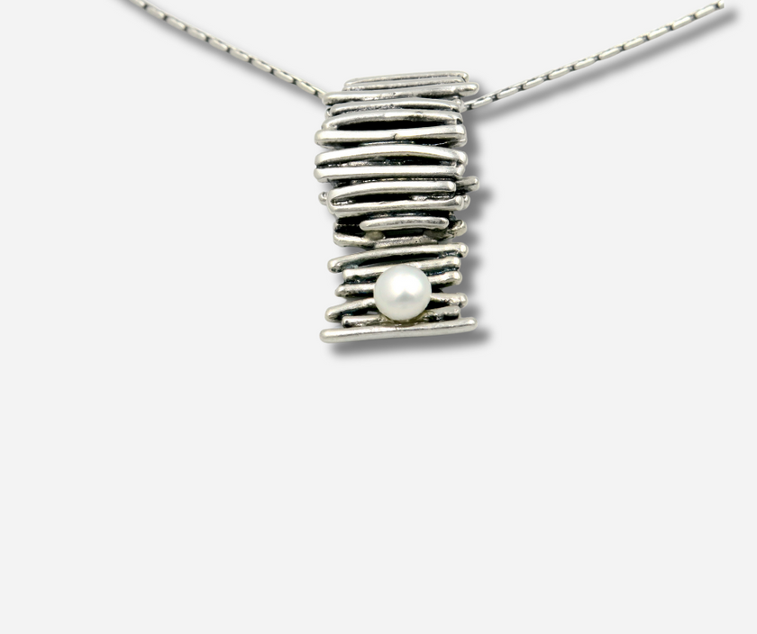 שרשרת אלגנטית בסגנון מודרני-תעשייתי עם תליון כסף בצורת גליל מלופף – חוטי כסף דקים כרוכים זה בזה ליצירת מרקם תלת-ממדי. בתחתיתו של הגליל שובצה פנינה לבנה קטנה, שמוסיפה נגיעה קלאסית לניגוד בין מתכת לברק טבעי. כל הפריט מוקפד ומעוצב לשדר מינימליזם יוקרתי ואיזון בין גולמיות לרכות.