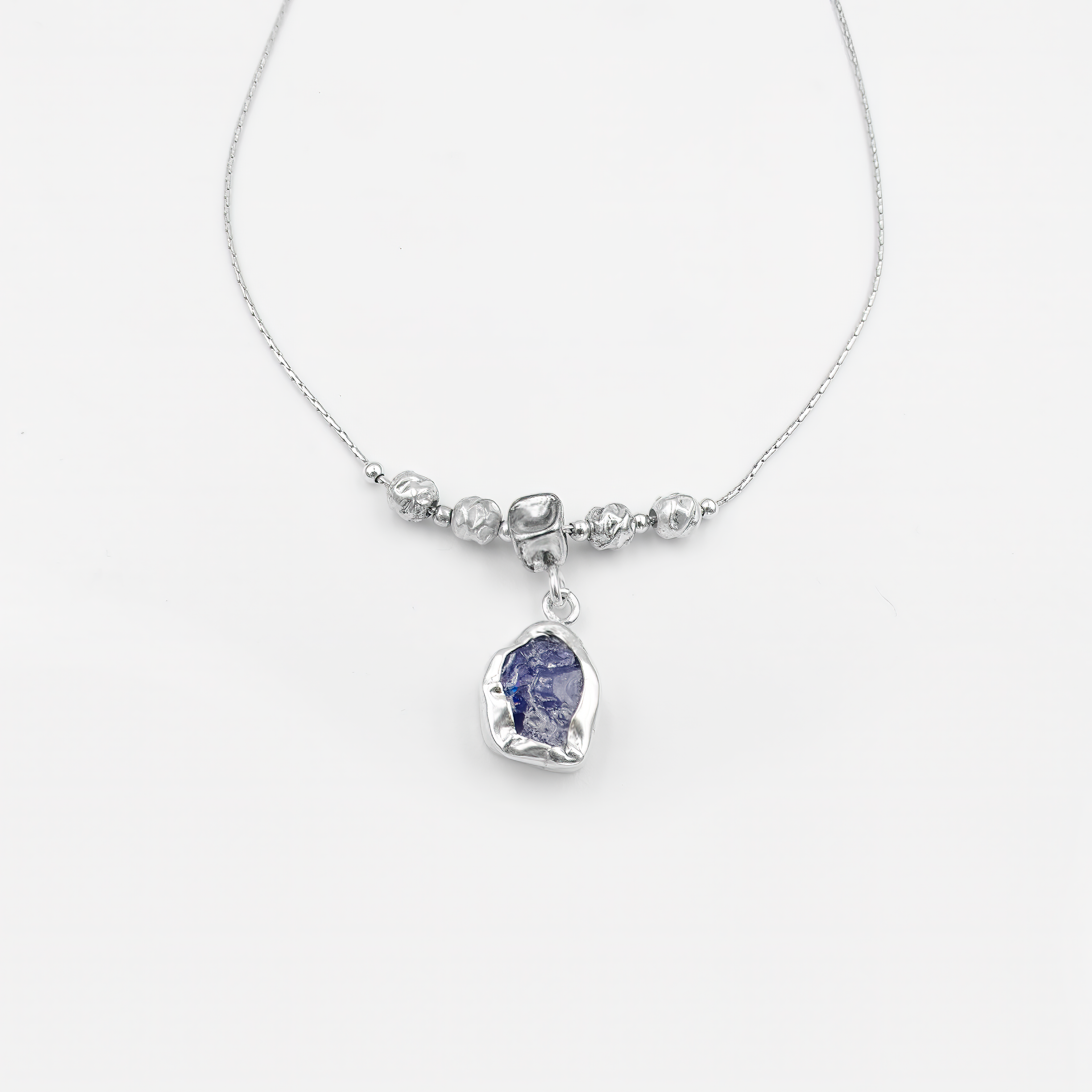 שרשרת מכסף סטרלינג בגימור מבריק, בעלת מרכז דקורטיבי הכולל חמישה חרוזים כסופים עגולים. תליון קטן ועגול משתלשל מתחת לחרוז המרכזי ומשובץ באבן כחולה בגוון קריסטלי-טבעי. האבן מזכירה את הגוון של קיאניט או ספיר לייט ומוסיפה אלמנט של עומק וצבע קריר. התליון במסגרת סגורה וקווים נקיים משתלב בצורה מושלמת עם שאר התכשיט, ומשדר איזון בין תחכום לפשטות.

