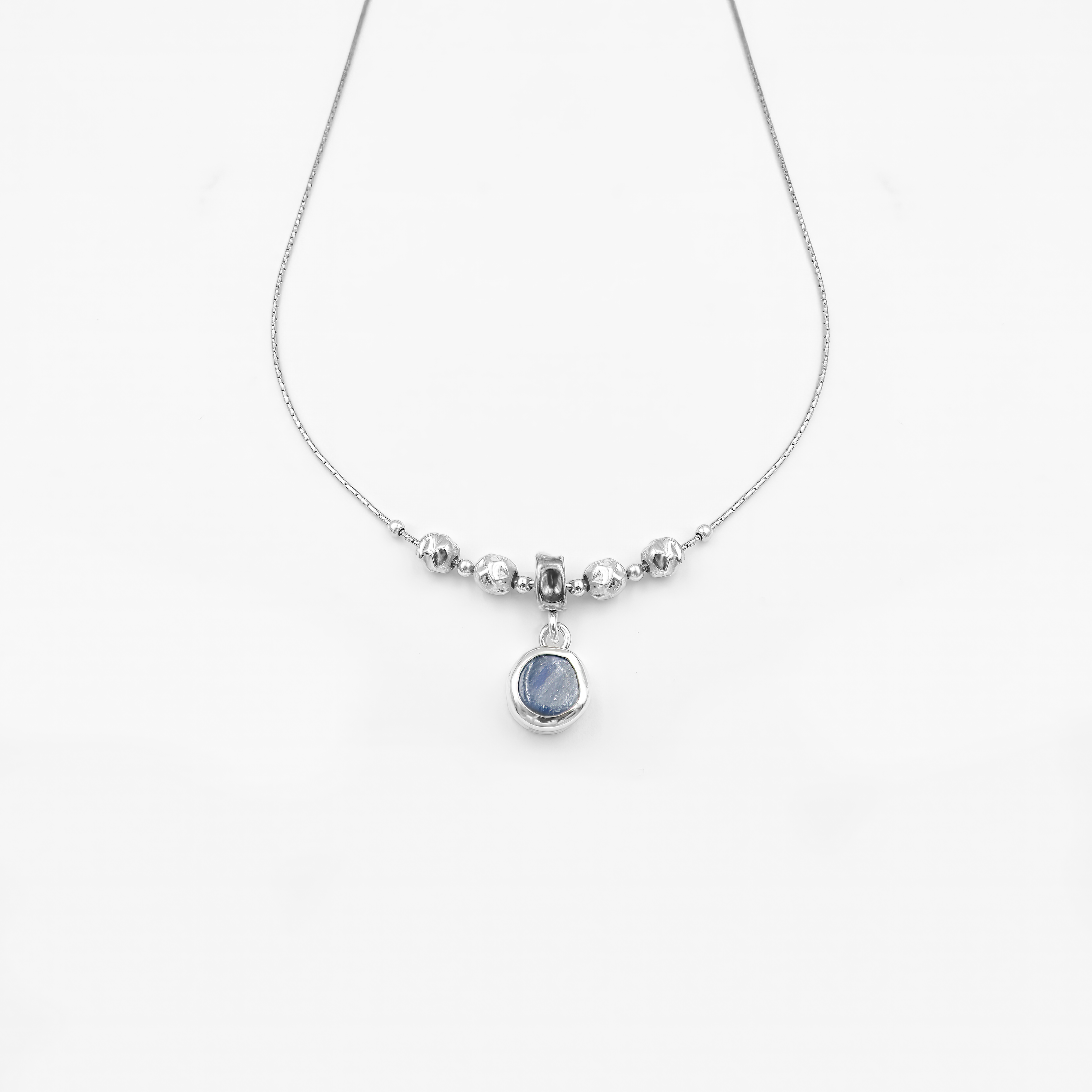 שרשרת אלגנטית בעיצוב מינימליסטי ומעודן, עשויה כסף מבריק עם שרשרת דקה ועדינה. במרכז השרשרת שורת חרוזים כסופים מעוגלים, היוצרים איזון סימטרי, ומתחתיהם תלוי תליון עגול קטן המשובץ באבן חן כחולה בגוון שמיים-עמוק. האבן בעלת מרקם טבעי ופנימי משתנה, מה שמוסיף לתכשיט נוכחות רגועה אך בולטת. מתאים למראה יומיומי עם טאץ' אלגנטי או כלוק ערב שקט ומעודן.