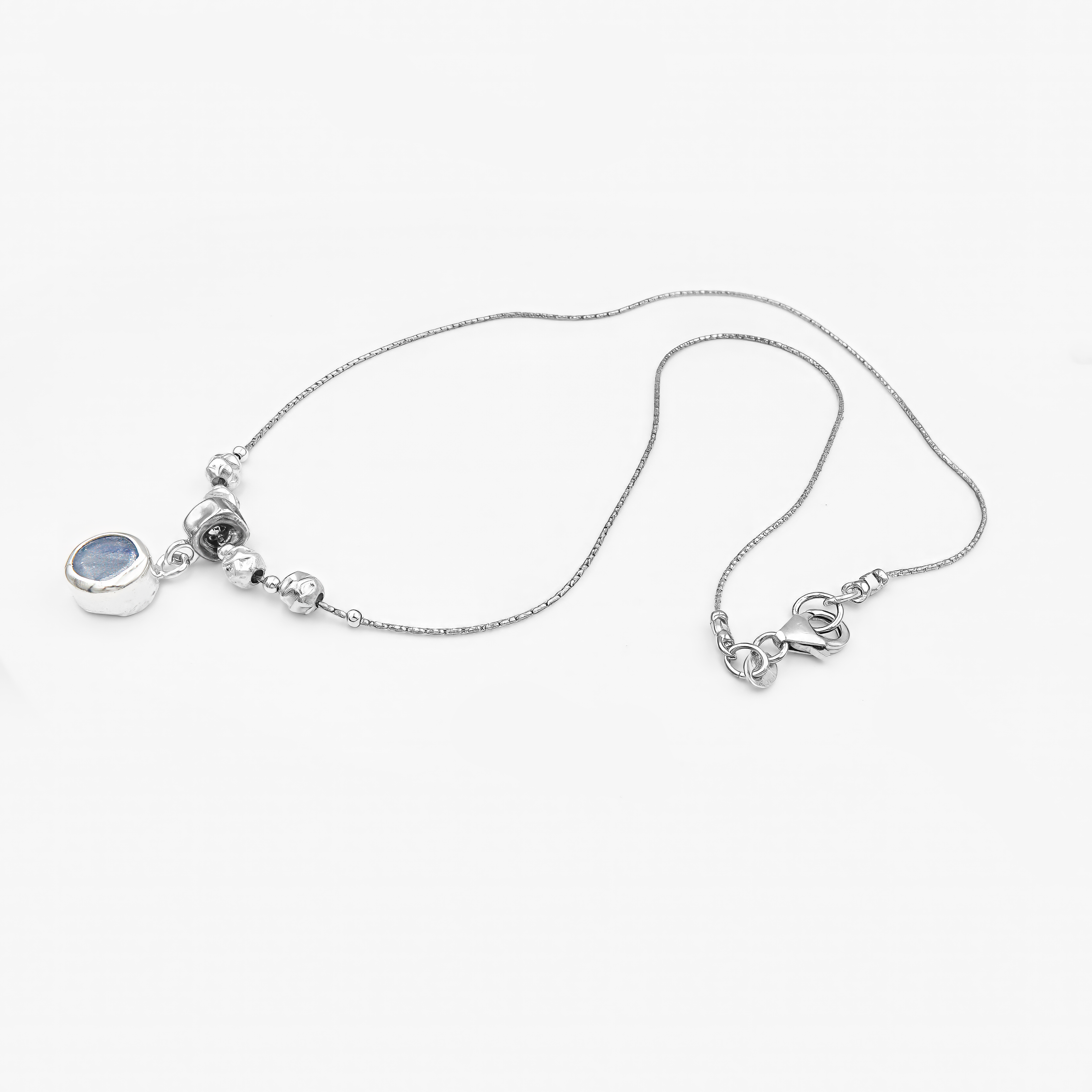 שרשרת מכסף סטרלינג בגימור מבריק, בעלת מרכז דקורטיבי הכולל חמישה חרוזים כסופים עגולים. תליון קטן ועגול משתלשל מתחת לחרוז המרכזי ומשובץ באבן כחולה בגוון קריסטלי-טבעי. האבן מזכירה את הגוון של קיאניט או ספיר לייט ומוסיפה אלמנט של עומק וצבע קריר. התליון במסגרת סגורה וקווים נקיים משתלב בצורה מושלמת עם שאר התכשיט, ומשדר איזון בין תחכום לפשטות.

