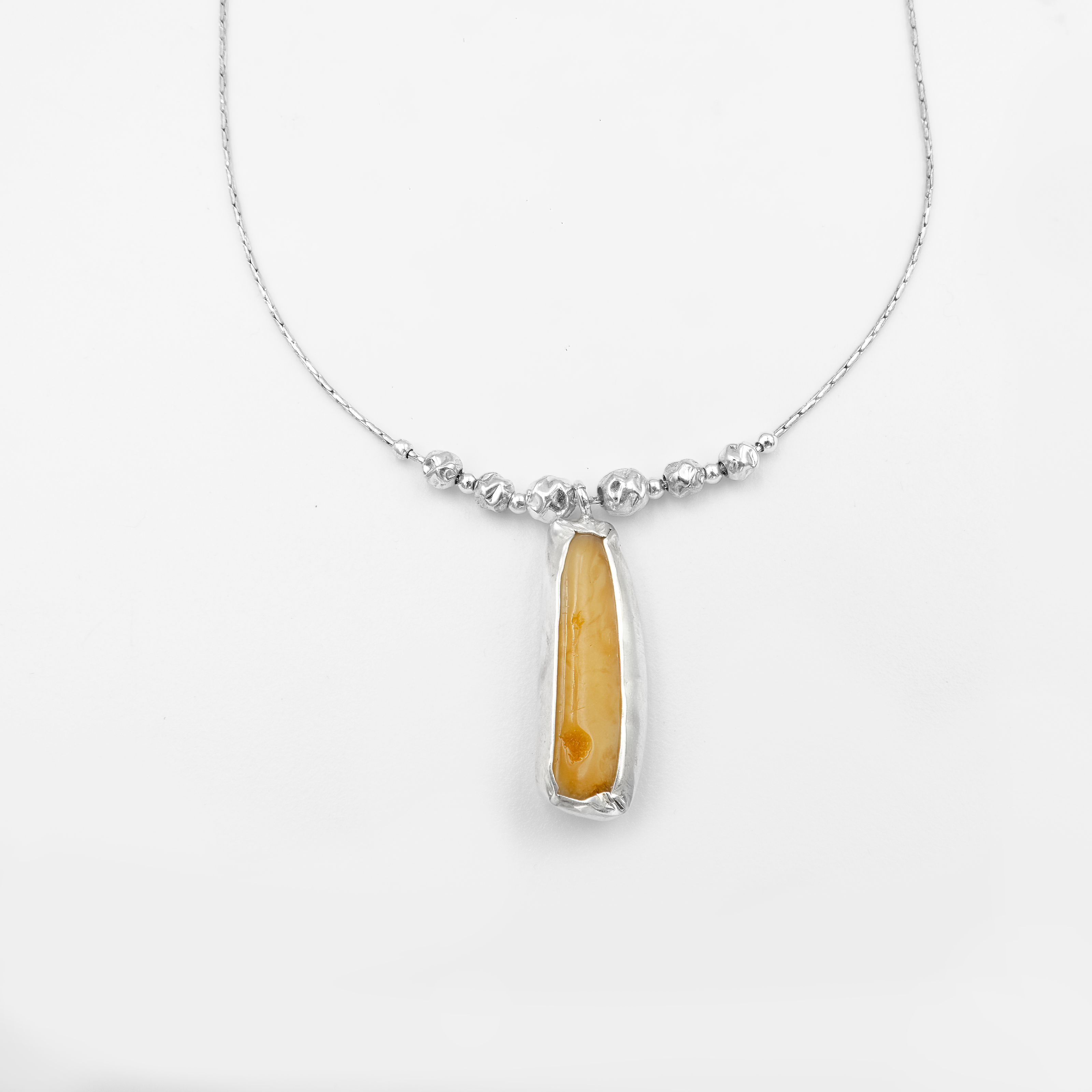 שרשרת כסף אלגנטית עם שורת חרוזים כסופים מבריקים במרכזה, ומתחתיהם תליון עגול קטן המשובץ באבן ענבר טבעית. גוון הענבר חמים – בין זהוב לכתום עם נגיעות של שקיפות, וייתכן שנראים בתוכה הכלואים הטבעיים האופייניים לאבן כמו בועות או סיבים עתיקים. מסגרת התליון עשויה כסף חלק, מה שמדגיש את צבעי הענבר ומבליט את עומקה. עיצוב שמשלב קווים נקיים עם נגיעה של היסטוריה טבעית, חמימות וזוהר רך.