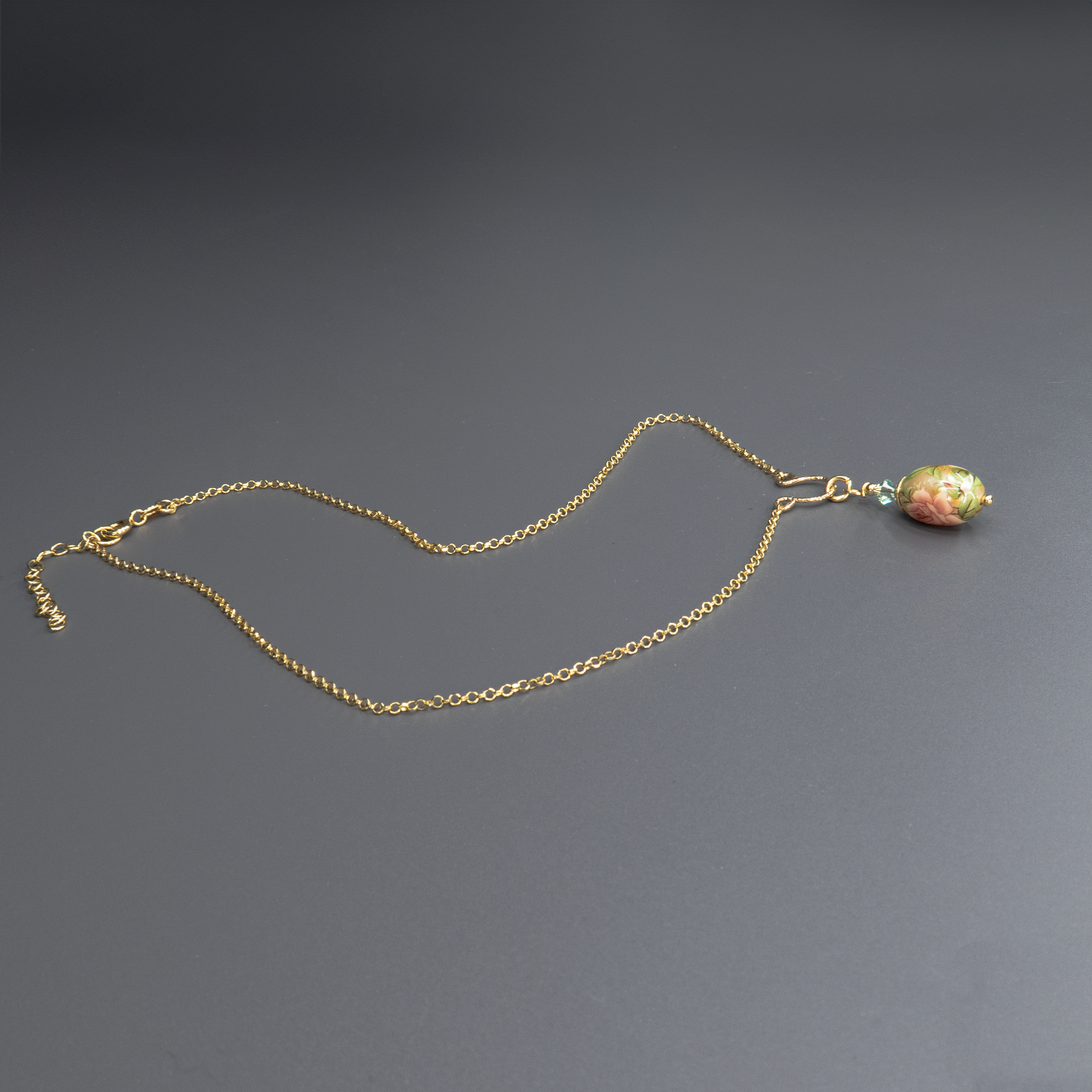 צילום של שרשרת מוזהבת עם תליון ביצה מצויר בעבודת יד, עם פרח בגווני ירוק מעושן, עלים בצבע זית כהה ודגש על קווים עדינים בגווני זהב וכחול. החרוזים המחברים בצבע ירוק כהה משדרים עומק ואלגנטיות. תכשיט מלא אופי ותחושת שלווה.