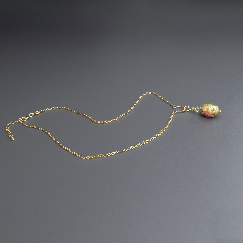 צילום של שרשרת מוזהבת עם תליון ביצה מצויר בעבודת יד, עם פרח בגווני ירוק מעושן, עלים בצבע זית כהה ודגש על קווים עדינים בגווני זהב וכחול. החרוזים המחברים בצבע ירוק כהה משדרים עומק ואלגנטיות. תכשיט מלא אופי ותחושת שלווה.