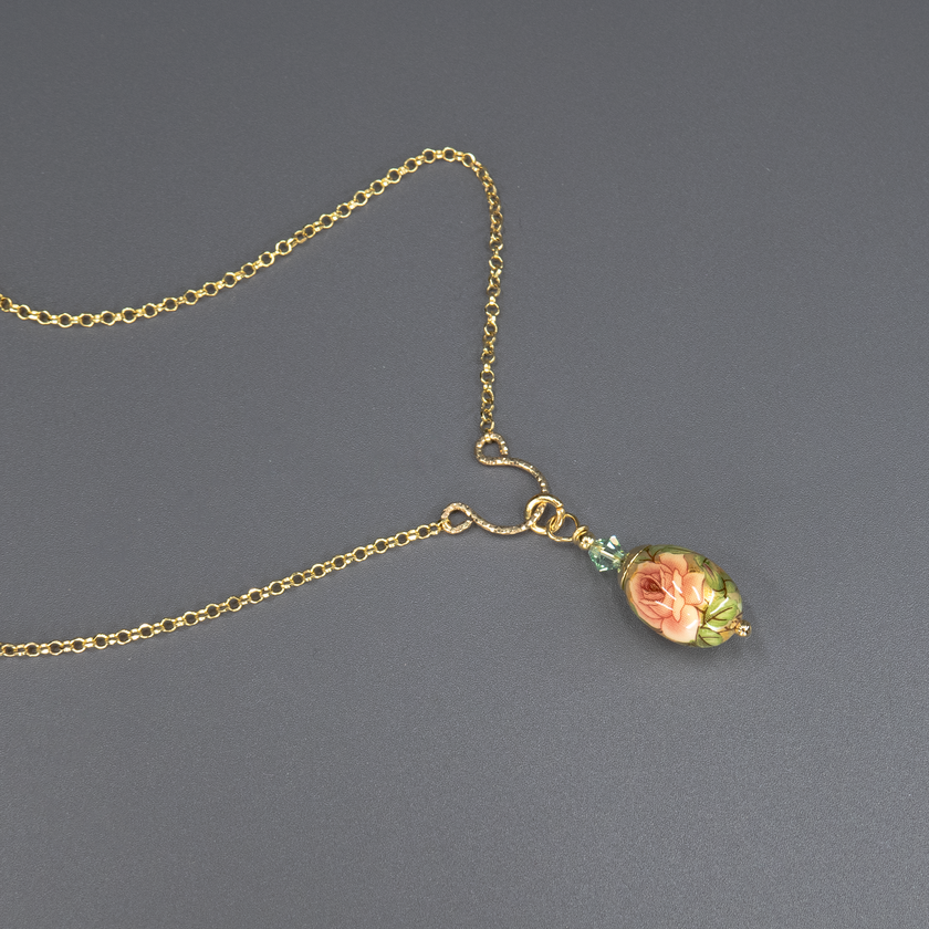 שרשרת אלגנטית בעיצוב וינטג' עם תליון קרמי אובלי בגוון ירקרק, מצויר ביד עם פרח רך בגווני ירוק בהיר, מוקף עלים כהים ופרחים זעירים. החרוזים המחברים בולטים בגוון ירוק אזמרגד מבריק. תכשיט שמזכיר יער אביבי וטבע רגוע.