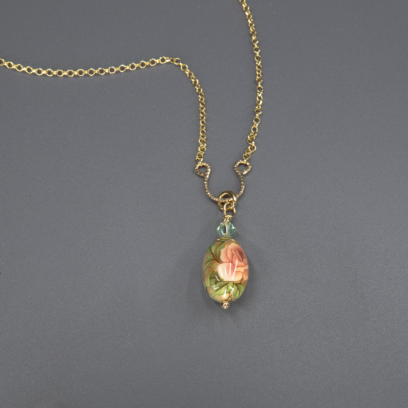 תכשיט בעבודת יד הכולל שרשרת מצופה זהב ותליון ביצה מעוצבת עם ציור פרח בגוון ירוק-מנטה, על רקע בהיר. העלים מסביב בגוונים של ירוק כהה וירוק זית, עם נגיעות כחולות עדינות. תכשיט רומנטי ומעודן בסגנון גינה אנגלית קסומה.