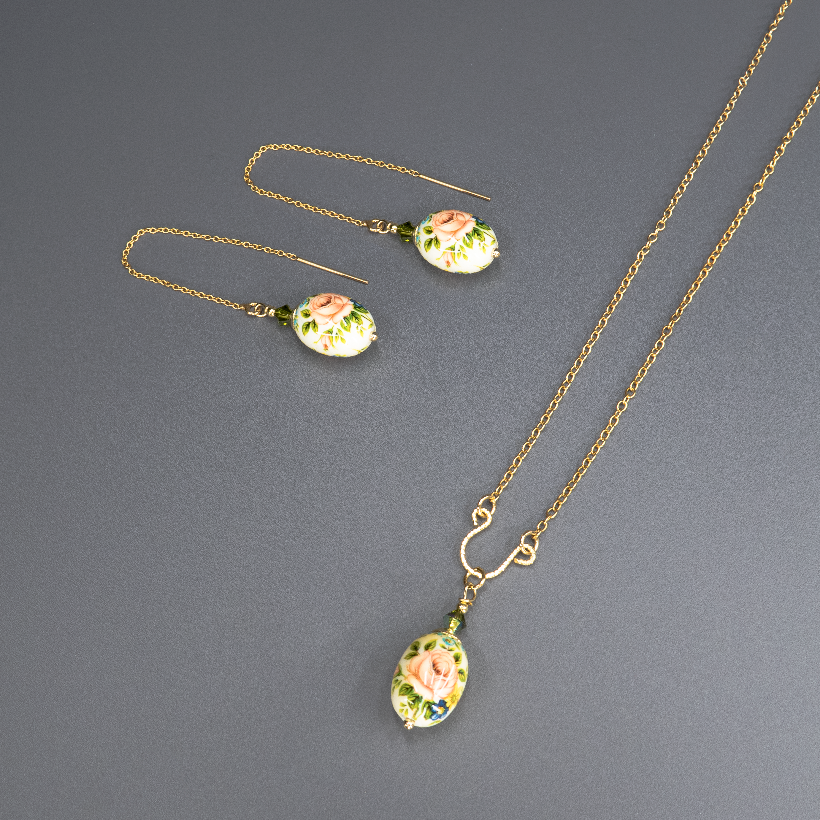 שרשרת אלגנטית מצופה זהב עם תליון אובלי דמוי ביצה, עשוי חומר קרמי או זכוכית מצוירת ביד, בגווני ורוד, ירוק וכחול. במרכז התליון מצויר ורד עדין בגוון אפרסק-ורוד, מוקף בעלים ירוקים ופרחי שדה קטנים בכחול. התליון תלוי בעדינות מחרוזים ירקרקים שמוסיפים עומק וזוהר. תכשיט בסגנון רומנטי וינטג', המשלב אומנות מסורתית עם ניחוח אירופאי עתיק.
העגיעלים להמחשה בלבד