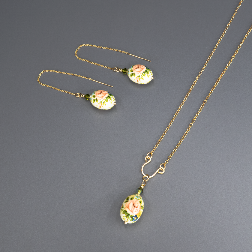 שרשרת אלגנטית מצופה זהב עם תליון אובלי דמוי ביצה, עשוי חומר קרמי או זכוכית מצוירת ביד, בגווני ורוד, ירוק וכחול. במרכז התליון מצויר ורד עדין בגוון אפרסק-ורוד, מוקף בעלים ירוקים ופרחי שדה קטנים בכחול. התליון תלוי בעדינות מחרוזים ירקרקים שמוסיפים עומק וזוהר. תכשיט בסגנון רומנטי וינטג', המשלב אומנות מסורתית עם ניחוח אירופאי עתיק.
העגיעלים להמחשה בלבד