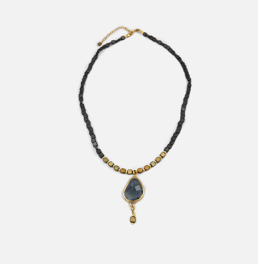 שרשרת מעוצבת בקווים אתניים עכשוויים, בשילוב חרוזים מרובעים שחורים וזהובים שמעניקים לה טקסטורה מעניינת. תליון האבן המרכזי מעוצב בצורת טיפה עם ליטוש שמדגיש את שקיפותו ואופיו הפנימי של החומר – בגוון כחול-אפור עמוק. המסגרת המוזהבת שלו מוסיפה חום וזוהר. בתחתיתו תלויה יחידת חרוז מרובעת נוספת בזהב ושחור, שמעניקה תחושת המשכיות ואסתטיקה מחושבת. שילוב מושלם בין מודרניות לחומרי גלם טבעיים.