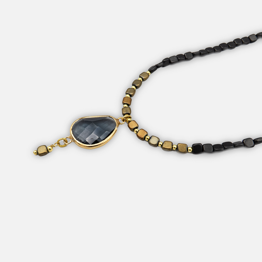 שרשרת גיאומטרית מיוחדת שמשלבת עיצוב אלגנטי עם חומרי גלם בעלי אופי. אבן מרכזית בגוון אפור כהה עם שקיפות קלה, מעוצבת בצורת טיפה ומשובצת במסגרת זהב מבריק. האבן משתלבת בהרמוניה עם חרוזים מרובעים קטנים בגווני שחור, זהב ונחושת. בתחתית התליון משתלשל חרוז מרובע נוסף שמשלים את הקומפוזיציה. העיצוב כולו משדר עידון, תחכום ואהבה לפרטים הקטנים – מושלם כפריט ייחודי למראה ערב או יומיומי מיוחד.