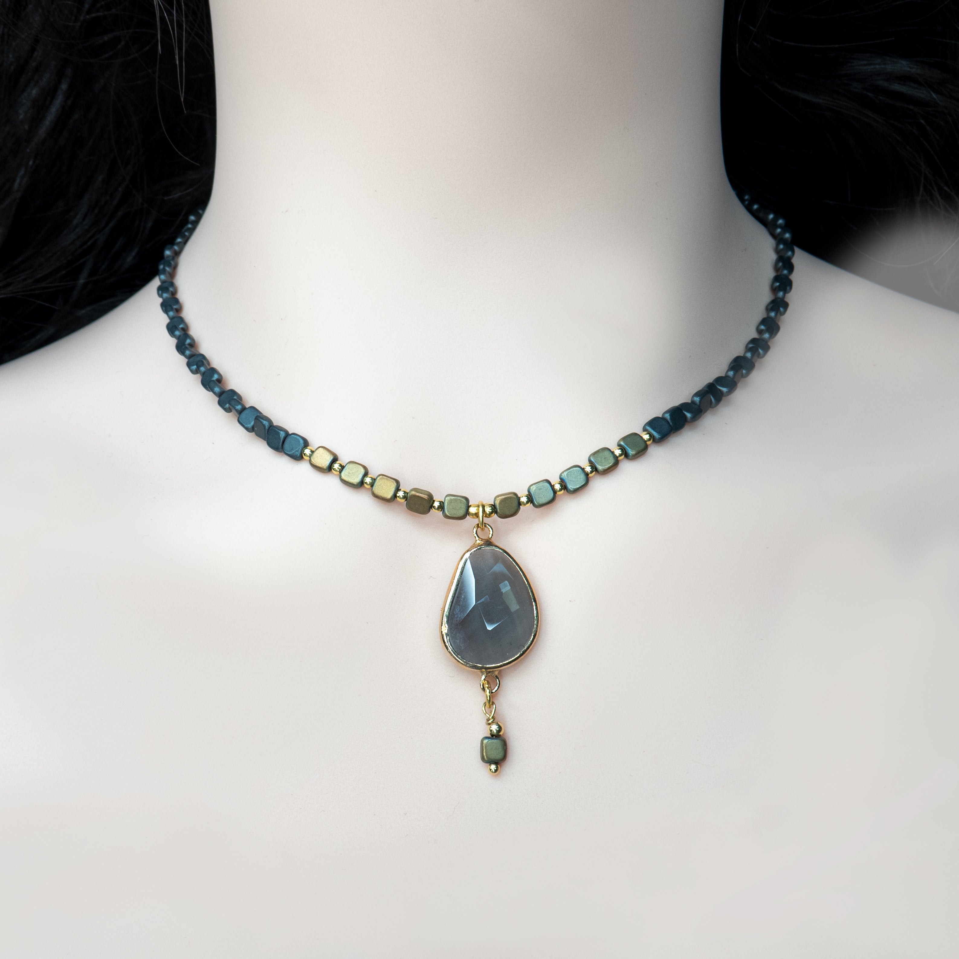 תכשיט נשי ומרשים עם אופי אומנותי: שרשרת קצרה עם חרוזים מרובעים בגווני שחור וזהב מטאליים, משתלבים בדפוס הרמוני לאורך כל ההיקף. במרכזה, תליון בצורת טיפה עשוי אבן אפורה כהה בעלת שקיפות עדינה וליטוש שטוח, מוקף במסגרת מוזהבת בגימור מבריק. מהתליון משתלשלת יחידת חרוז נוספת, שיוצרת תחושת תנועה ועומק. השרשרת משדרת איזון בין נוכחות חזקה לאלגנטיות עדינה – תכשיט בעל נראות על-זמנית שמתאים ליום ולערב.