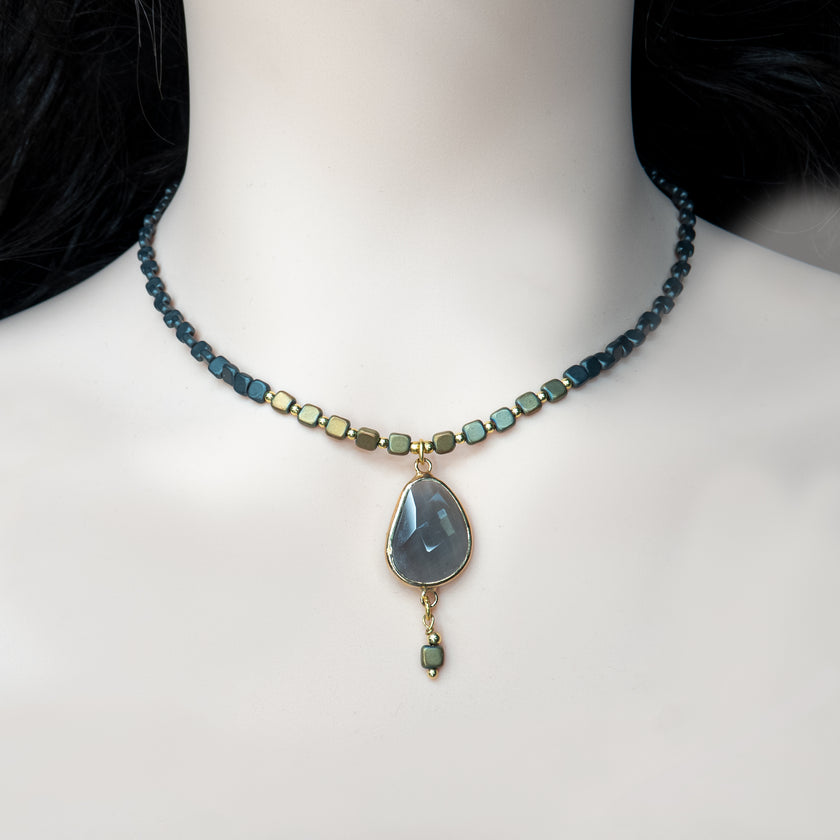 תכשיט נשי ומרשים עם אופי אומנותי: שרשרת קצרה עם חרוזים מרובעים בגווני שחור וזהב מטאליים, משתלבים בדפוס הרמוני לאורך כל ההיקף. במרכזה, תליון בצורת טיפה עשוי אבן אפורה כהה בעלת שקיפות עדינה וליטוש שטוח, מוקף במסגרת מוזהבת בגימור מבריק. מהתליון משתלשלת יחידת חרוז נוספת, שיוצרת תחושת תנועה ועומק. השרשרת משדרת איזון בין נוכחות חזקה לאלגנטיות עדינה – תכשיט בעל נראות על-זמנית שמתאים ליום ולערב.