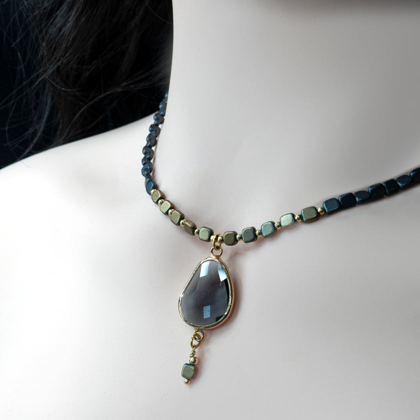 שרשרת קלאסית עם טוויסט עכשווי – מחרוזים מרובעים שחורים בגימור מט, משולבים עם חרוזים קטנים בגוון זהב עתיק ומתכתי. במרכזה תליון בצורה אורגנית של טיפה, עשוי אבן מלוטשת בגוון כהה עם עומק פנימי מהפנט, משובץ במסגרת מוזהבת. לתליון מחובר חרוז נוסף שתלוי חופשי, מוסיף תנועה רכה ונגיעה אמנותית. תכשיט שמתאים לאוהבות סגנון בוהו-שיק יוקרתי, עם נוכחות שקטה אך בלתי נשכחת.

