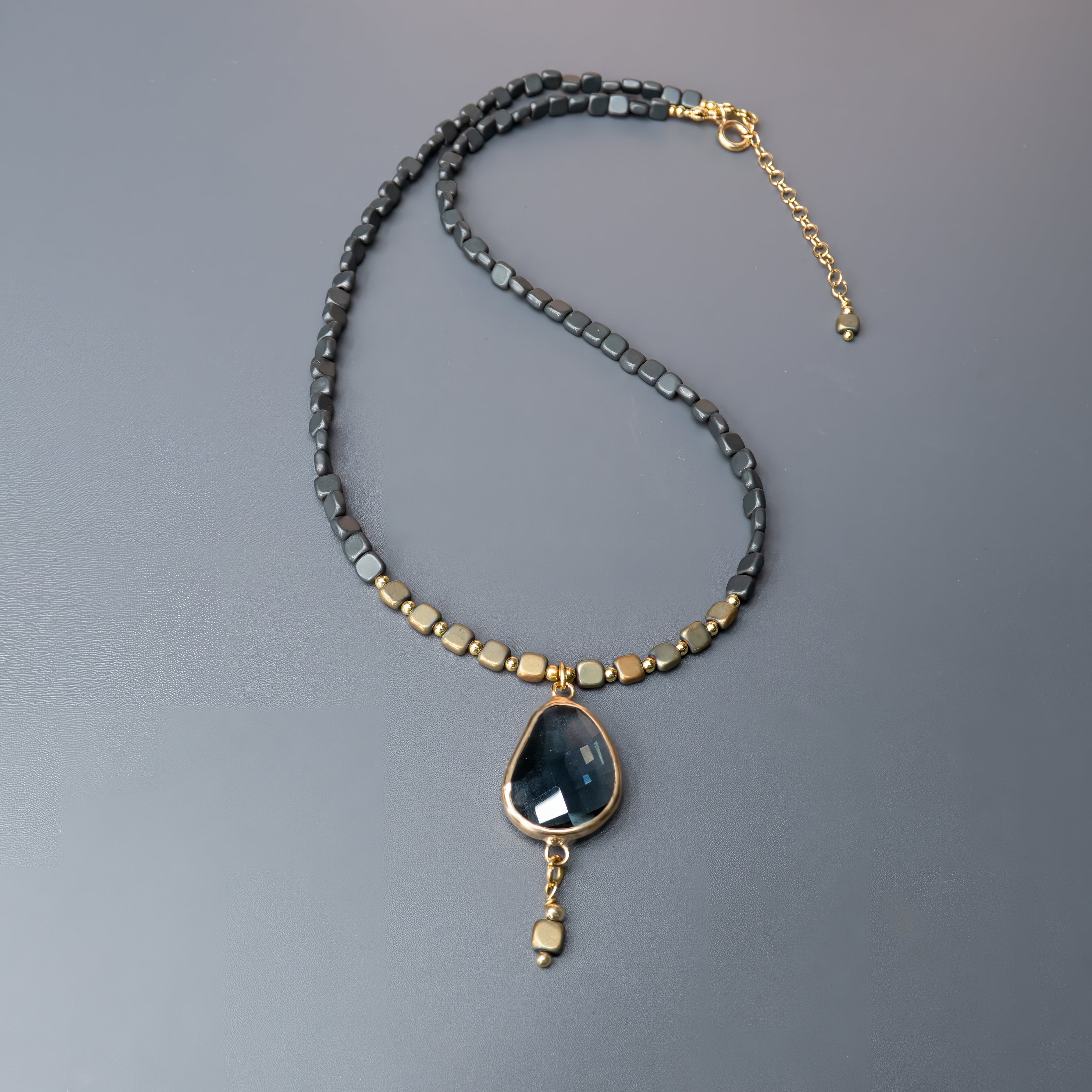 תכשיט בעל נוכחות עזה ומראה עוצמתי – שרשרת עם חרוזים מרובעים קטנים בגווני שחור מתכתי וזהב עתיק, שמקנים לה טקסטורה עשירה. במרכזה תליון טיפה גדול עם אבן חצי-שקופה בגוון גרפיט, בעלת ליטוש עדין ומראה פנימי מסתורי. התליון ממוסגר בזהב בוהק, ומהתחתית משתלשל חרוז נוסף שמדגיש את האורך והאלגנטיות. שרשרת שמדברת בשפה של עיצוב מודרני ונגיעה קלאסית – לאישה שאוהבת תכשיטים עם משמעות