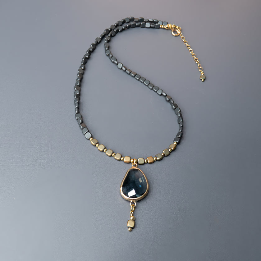 תכשיט בעל נוכחות עזה ומראה עוצמתי – שרשרת עם חרוזים מרובעים קטנים בגווני שחור מתכתי וזהב עתיק, שמקנים לה טקסטורה עשירה. במרכזה תליון טיפה גדול עם אבן חצי-שקופה בגוון גרפיט, בעלת ליטוש עדין ומראה פנימי מסתורי. התליון ממוסגר בזהב בוהק, ומהתחתית משתלשל חרוז נוסף שמדגיש את האורך והאלגנטיות. שרשרת שמדברת בשפה של עיצוב מודרני ונגיעה קלאסית – לאישה שאוהבת תכשיטים עם משמעות
