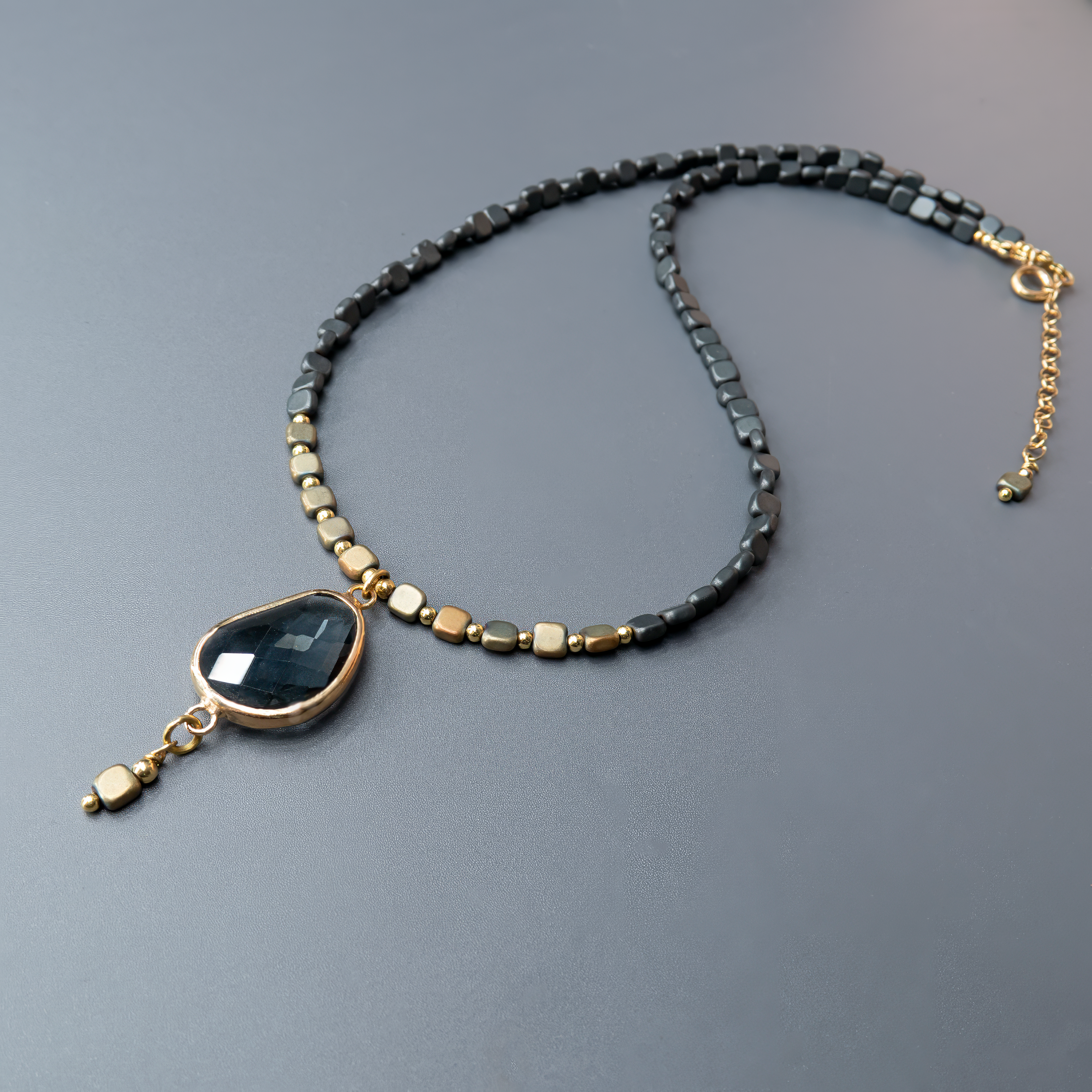 שרשרת קצרה בסגנון אתני עכשווי, עשויה משילוב של חרוזים מתכתיים כהים וזהובים, בתבנית משתנה ומקורית. במרכזה תליון טיפה מרהיב, עשוי אבן בגוון כהה עם ברק פנימי שמזכיר מים עמוקים או זכוכית עתיקה. האבן נתונה במסגרת מתכת זהובה, וממנה משתלשל חרוז מרובע קטן שמוסיף עניין חזותי ותנועה. עיצוב שמתחבר לרוח של מינימליזם עם טוויסט – פריט שימשוך מבטים.