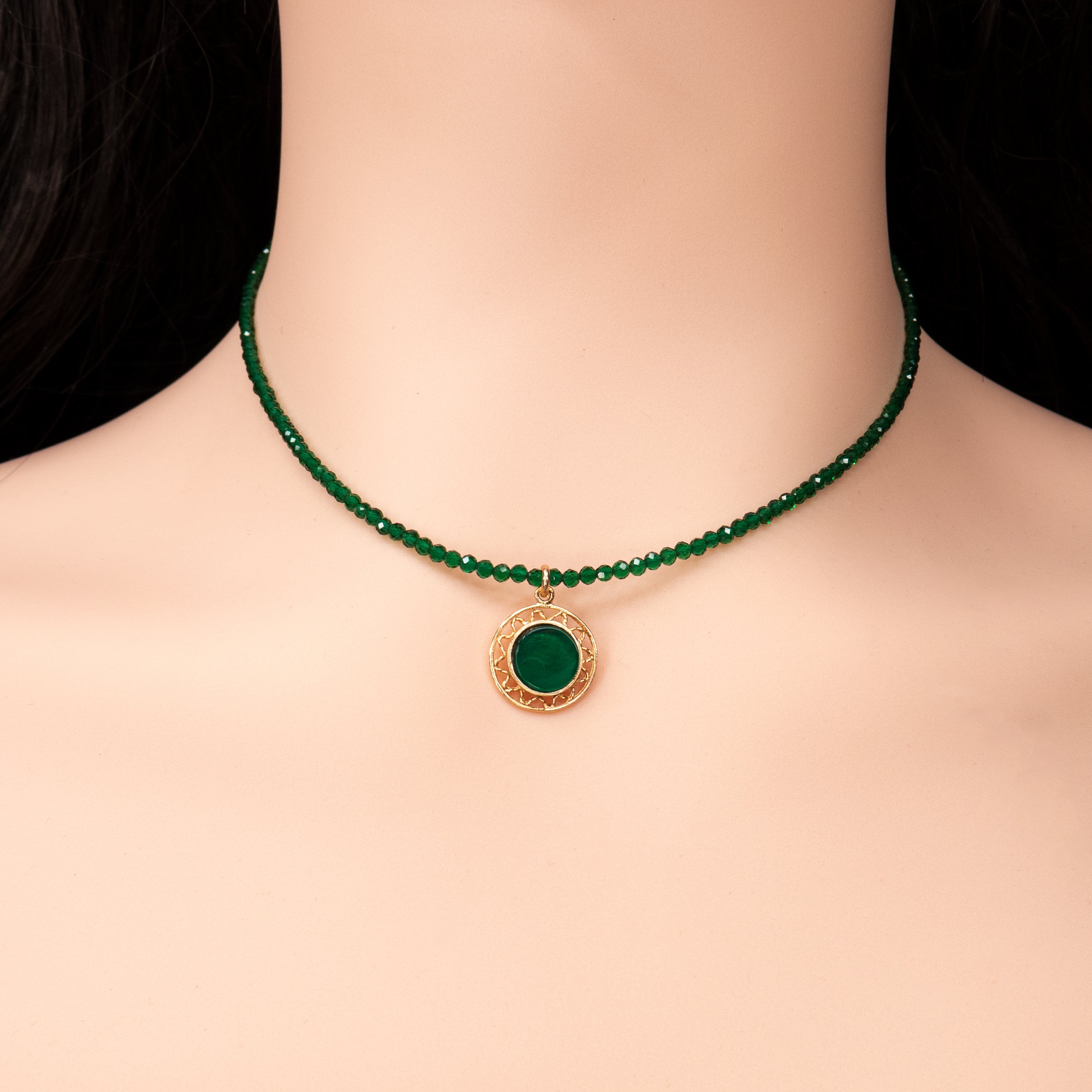 קולר בעבודת יד עשוי חרוזים ירוקים מבריקים בגודל אחיד, המסודרים על חוט גמיש בצמוד לצוואר. במרכזו תליון עגול מוזהב עם מסגרת חרוטה ואבן ירוקה משובצת במרכז. מונח על רקע של עור בהיר וחולצה לבנה מכופתרת. עיצוב מדויק שמשלב מראה אלגנטי עם נגיעה קלאסית.