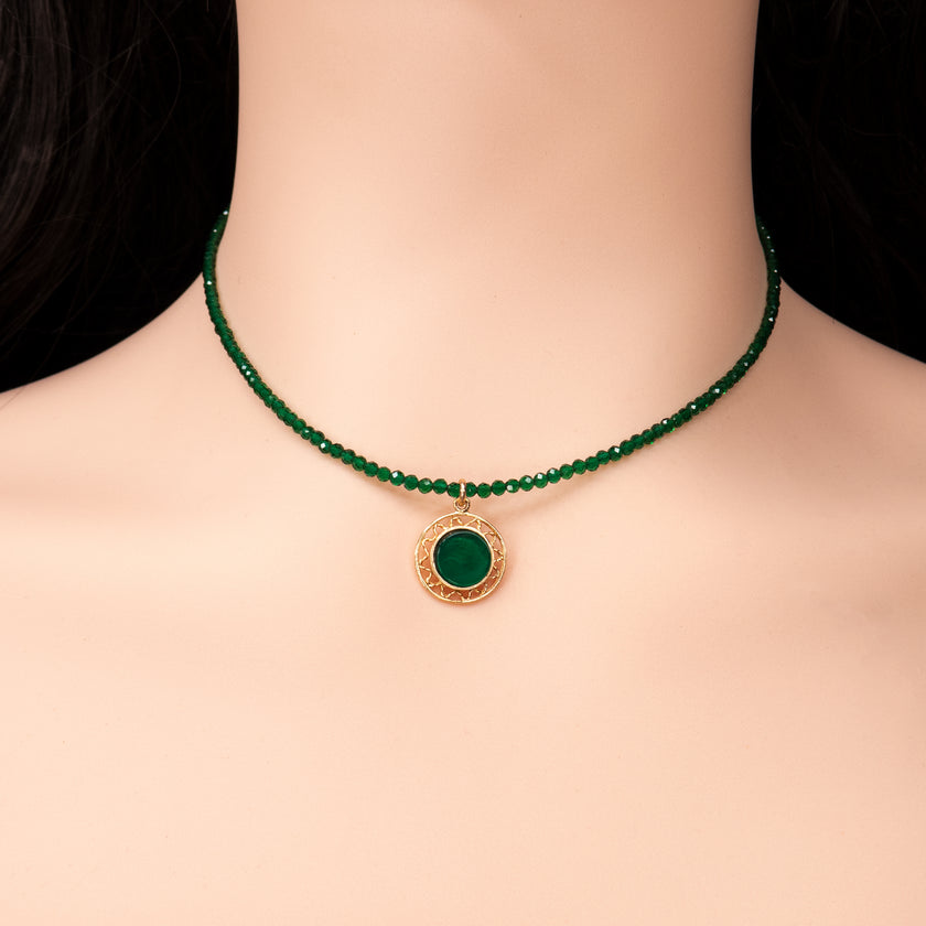 קולר בעבודת יד עשוי חרוזים ירוקים מבריקים בגודל אחיד, המסודרים על חוט גמיש בצמוד לצוואר. במרכזו תליון עגול מוזהב עם מסגרת חרוטה ואבן ירוקה משובצת במרכז. מונח על רקע של עור בהיר וחולצה לבנה מכופתרת. עיצוב מדויק שמשלב מראה אלגנטי עם נגיעה קלאסית.