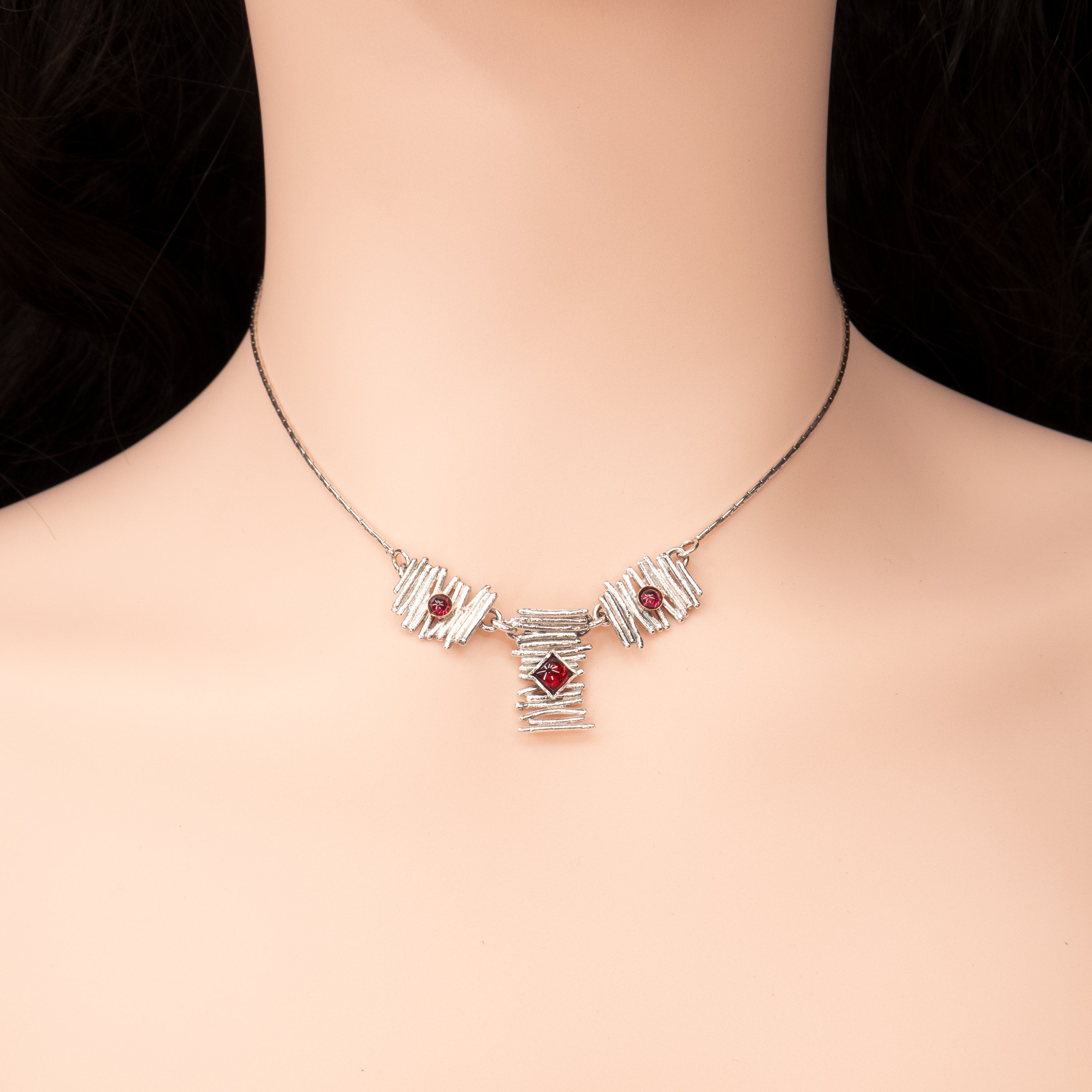 צילום תקריב של שרשרת כסף מודרנית על צוואר דוגמנית: שלושה אלמנטים טקסטורליים הבנויים מקווים דקים ולא-סימטריים. המרכזי ארוך ובמרכזו גרנט מרובע בגוון אדום-יין; בצדדים שני אלמנטים קטנים יותר עם גרנטים עגולים. השרשרת דקה ועדינה, יושבת מעל עצם הבריח, והרקע בגוון עור ניטרלי שמדגיש את הברק הכסוף ואת האודם של האבנים.