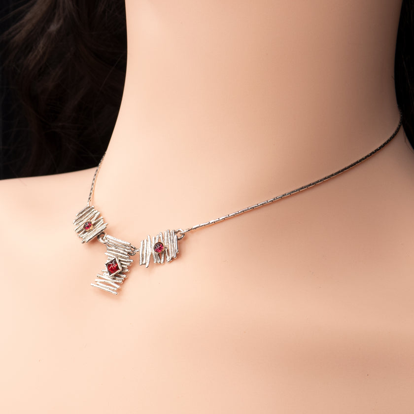 תכשיט קצר בעיצוב גאומטרי-אורגני: שלושה לוחות כסף המדמים ערימות ענפים עדינים, כל אחד משובץ גרנט בורדו נוצץ. במרכז לוח אנכי עם גרנט מרובע; משני צדיו לוחות קטנים עם אבני גרנט עגולות. קו השרשרת הנקי מונח על צוואר בהיר, ויוצר קונטרסט בין הברק הכסוף לצבע האדום-עמוק.
