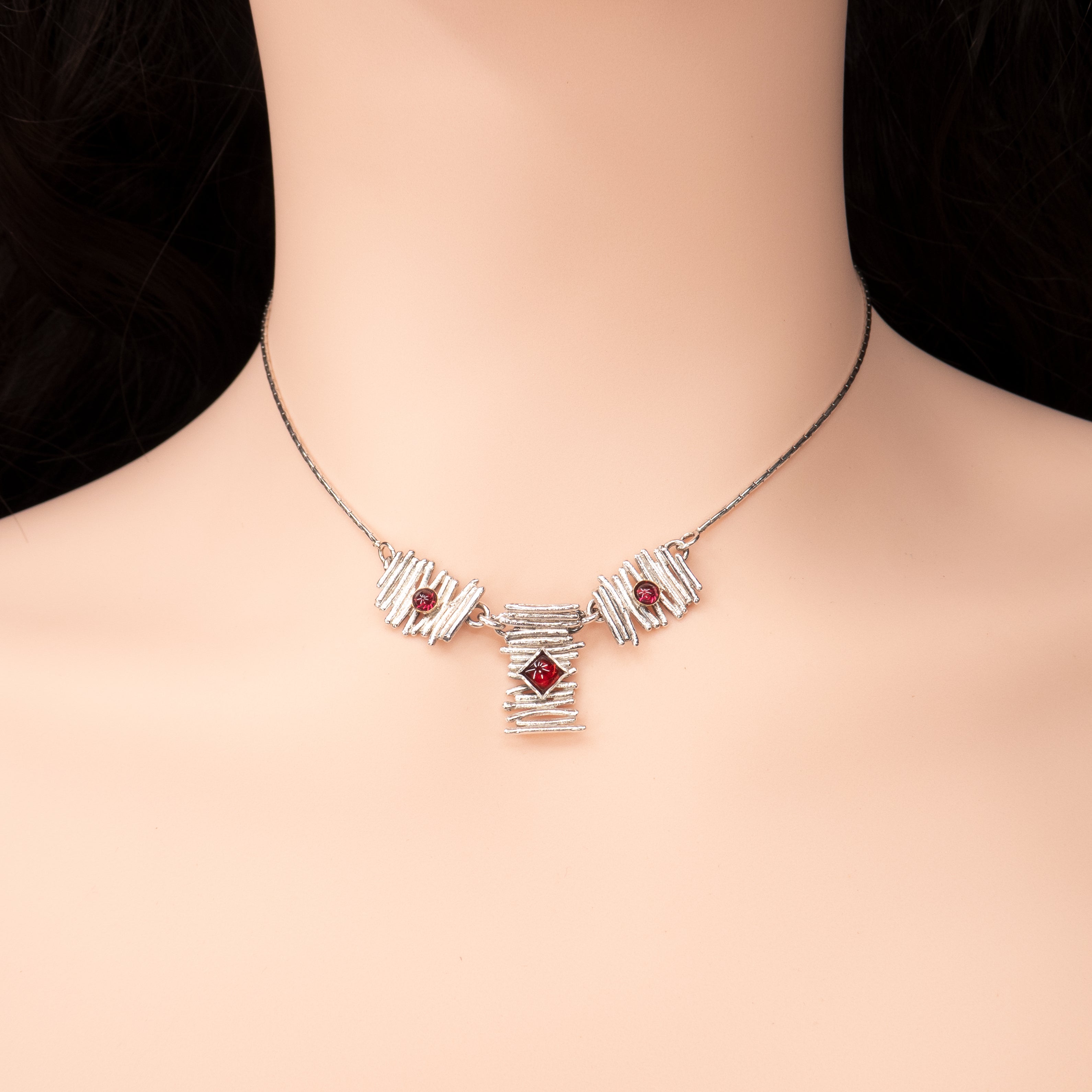 שרשרת כסף סטרלינג – מתנה מושלמת לבני ינואר, חודש הגרנט. שלושה רכיבים במרקם “מקלות” לא-סימטריים: ריבוע מרכזי משובץ גרנט מרובע ושני ריבועים צדדיים עם גרנטים עגולים. מוצג על צוואר חלק ורקע ניטרלי, המדגיש את הברק המתכתי ואת גוון האדום החושני של האבנים.