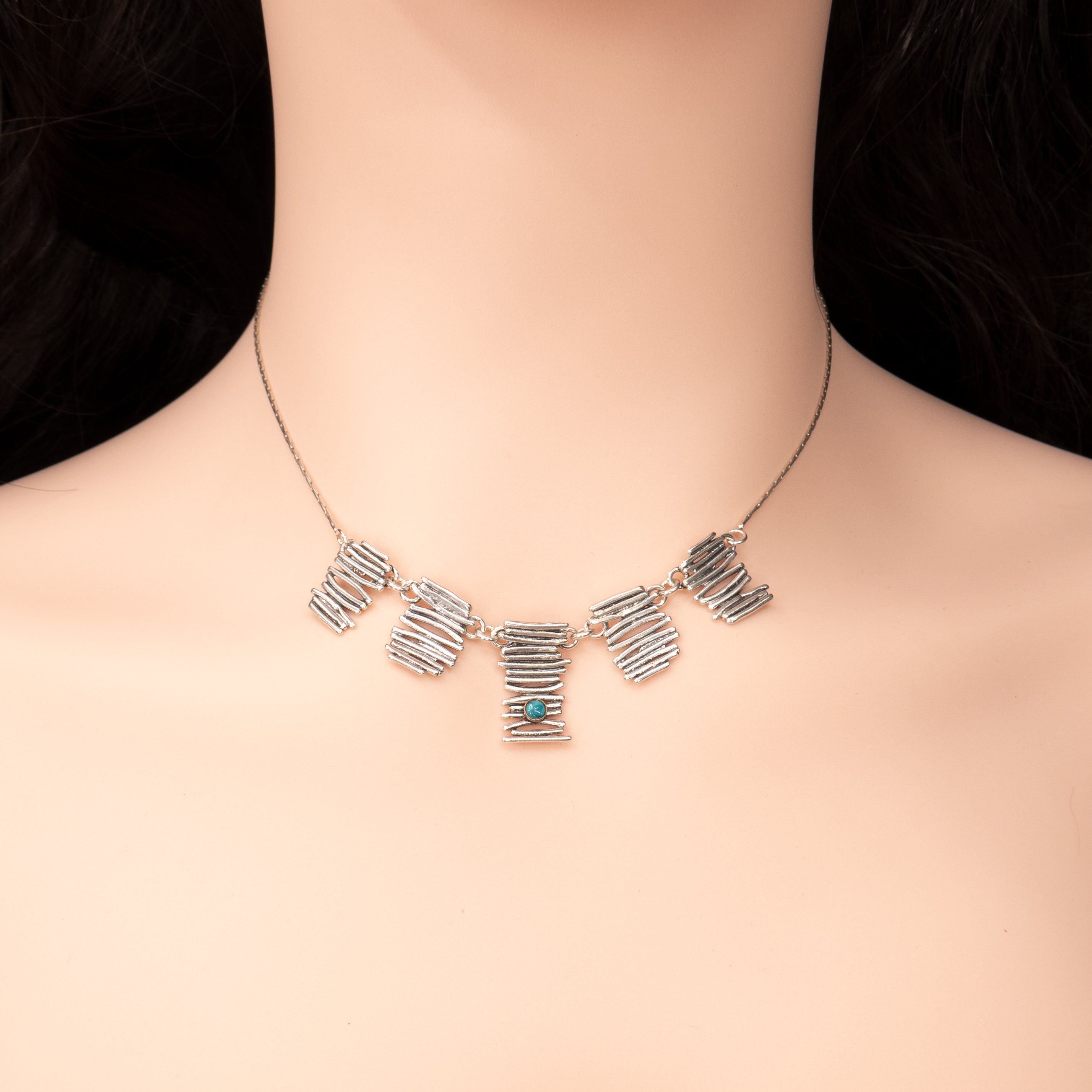 צילום תקריב של שרשרת כסף בעיצוב מודרני עם שלושה אלמנטים מלבניים, המעוצבים בטקסטורה גולמית המדמה ענפים או תבליטים. כל חלק משובץ באבן טורקיז בגוון כחול־שמיים עז – המרכזית בחיתוך מרובע, והצדדיות בחיתוך עגול. גוון הטורקיז הבוהק יוצר ניגוד מרהיב עם הכסף המוברש, ומעניק לתכשיט מראה חי, צעיר ובעל נוכחות.
