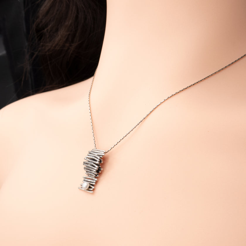 צילום של שרשרת כסף בסגנון מינימליסטי-אמנותי, עם תליון דמוי סליל מתכתי – חוטי כסף דקים המלופפים אחד על השני בצורה גלילית, יוצרים משחק מרקמים מעניין. בתחתית הסליל שובצה פנינה עגולה קטנה בגוון לבן פנינה, שמאזנת את העיצוב הגאומטרי עם נגיעה קלאסית ונשית. כל הפריט מוקפד, מדויק ומשדר טעם מודרני ונקי.