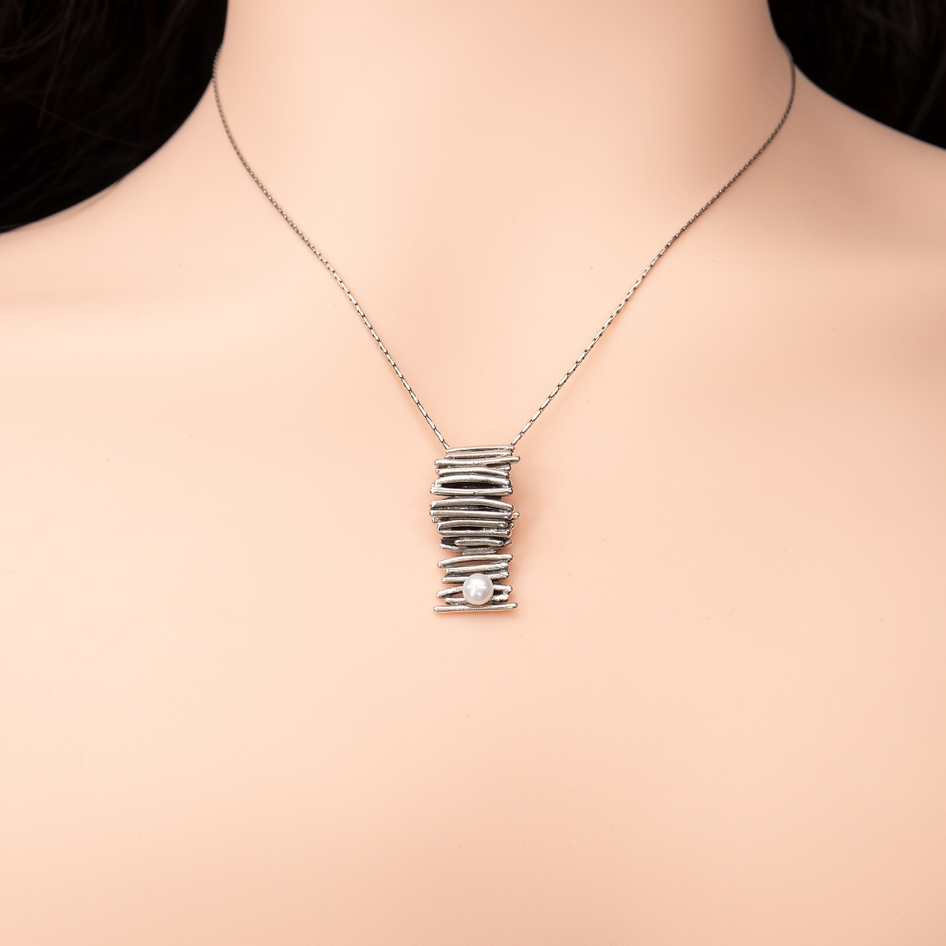תכשיט מינימליסטי ומדויק: שרשרת כסף סטרלינג דקה ונקייה, שבמרכזה תלוי תליון קומפקטי בעיצוב בלתי שגרתי – חוטי כסף שמלופפים בצפיפות סביב עצמם ויוצרים גליל ייחודי עם מרקם עשיר. בתחתית התליון משובצת פנינה קטנה בגוון שמנת. עיצוב שמתאים לאוהבות קווים נקיים, חומריות טבעית ומראה עכשווי.