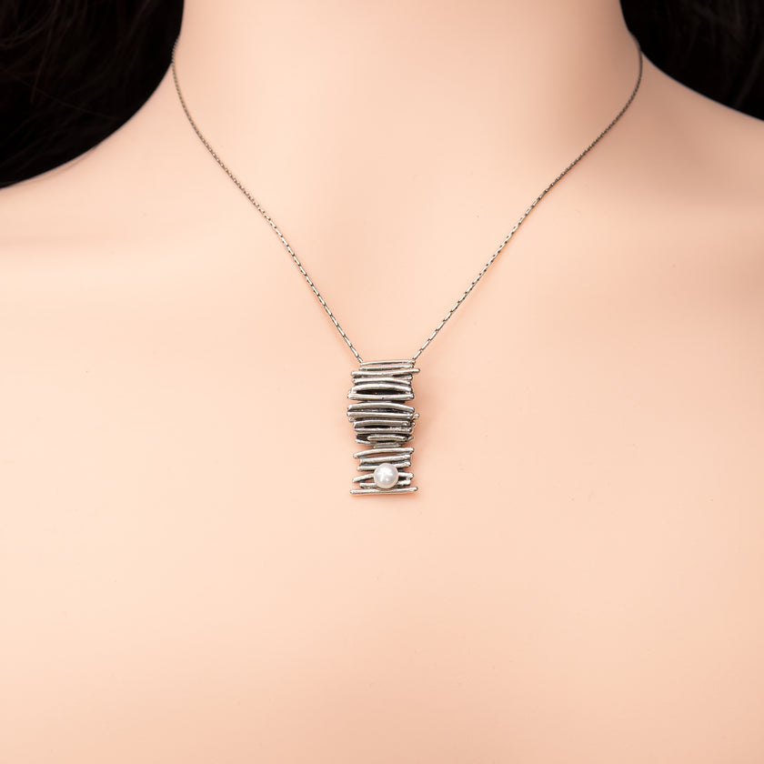 תכשיט מינימליסטי ומדויק: שרשרת כסף סטרלינג דקה ונקייה, שבמרכזה תלוי תליון קומפקטי בעיצוב בלתי שגרתי – חוטי כסף שמלופפים בצפיפות סביב עצמם ויוצרים גליל ייחודי עם מרקם עשיר. בתחתית התליון משובצת פנינה קטנה בגוון שמנת. עיצוב שמתאים לאוהבות קווים נקיים, חומריות טבעית ומראה עכשווי.