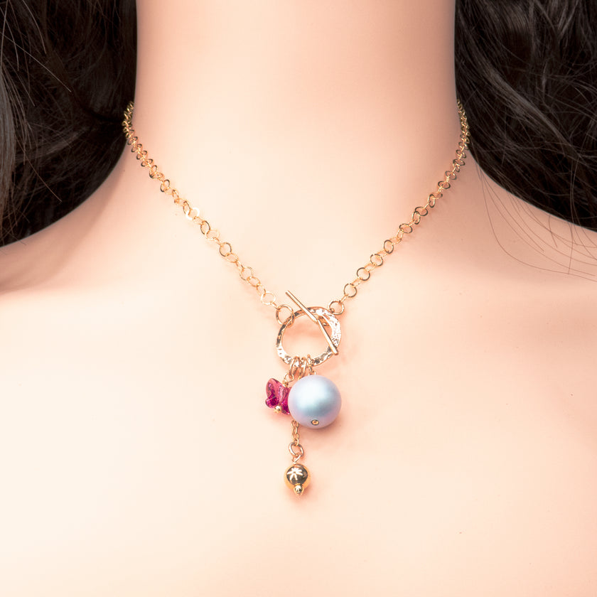 שרשרת עם סגירת טוגל קדמית בגוון זהב מבריק, תליון פנינה סגלגלה גדולה, חרוז בצורת פרפר בגוון ורוד עז, ותליון כדור זהוב. כל האלמנטים תלויים יחד ויוצרים תנועה ועניין. עיצוב שמחבר בין ילדותיות אלגנטית לקווים מודרניים.
