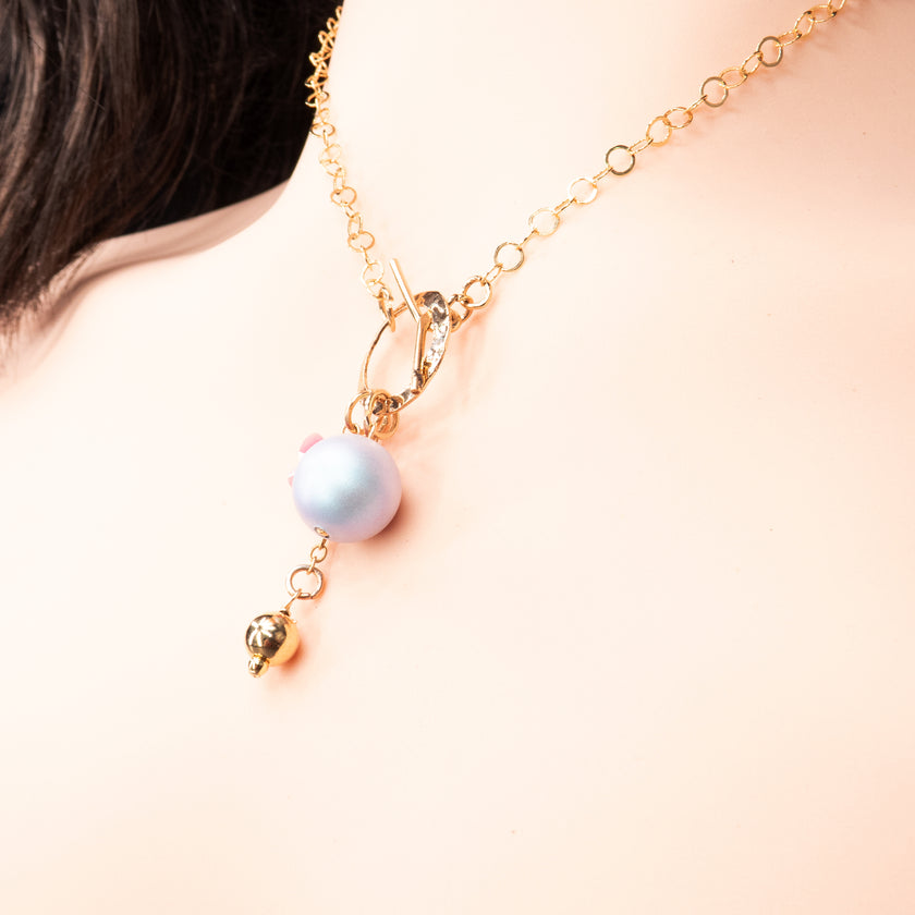 שרשרת מוזהבת עם עיצוב אסימטרי: פנינה בגוון כחול-סגלגל, פרפר זכוכית בגוון ורוד בוהק ותליון מתכתי קטן. כולם מחוברים לסוגר טוגל דקורטיבי קדמי, שמוסיף עניין עיצובי. השרשרת משדרת שמחה, נשיות וייחוד.