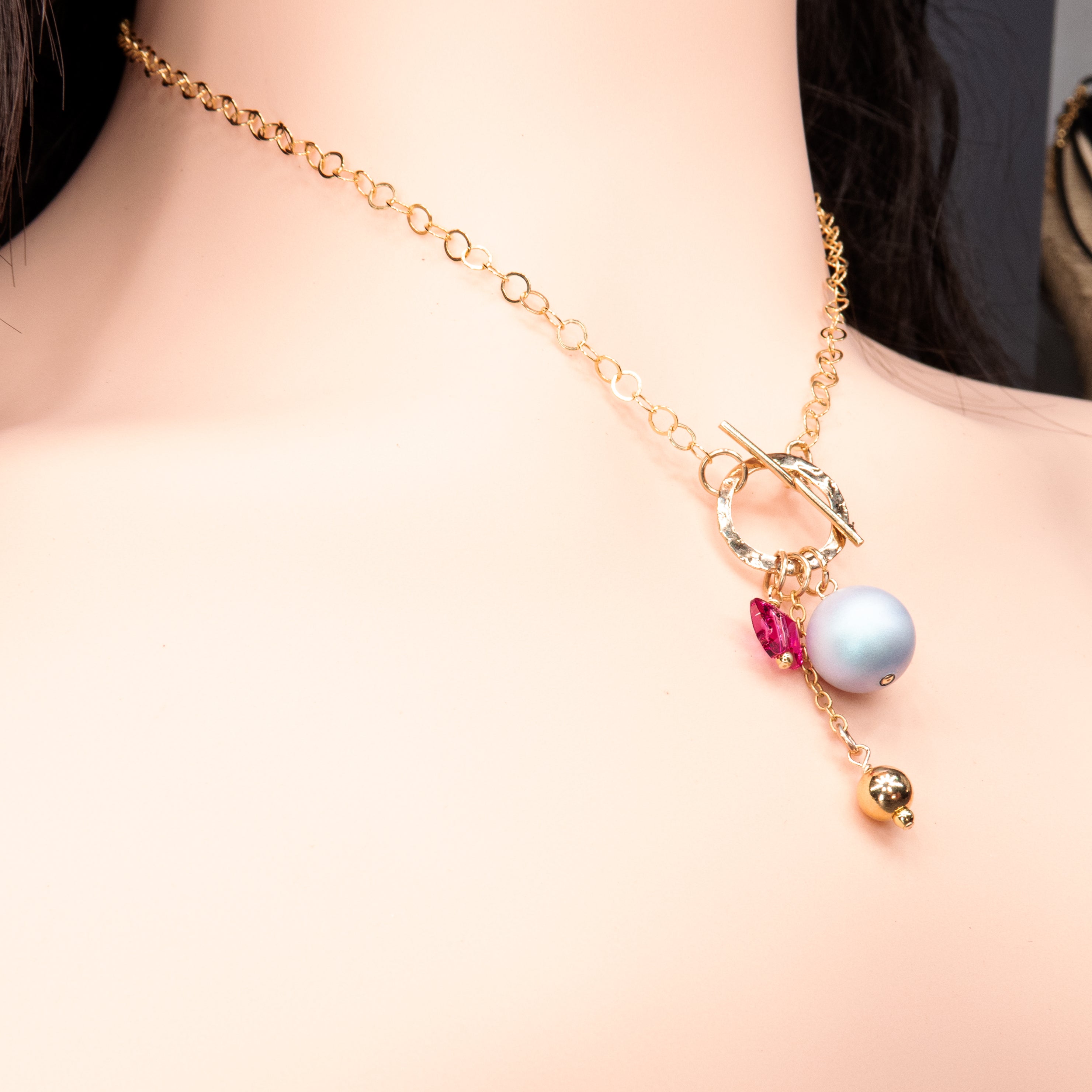 שרשרת בסגנון יוניק עם סוגר קדמי בגימור מטאלי מוזהב, תליון פנינה בגוון לילך, חרוז פרפר ורוד ותליון נוסף בזהב. כל האלמנטים תלויים יחד ויוצרים תכשיט נעים, מקורי ומלא שמחה צבעונית.