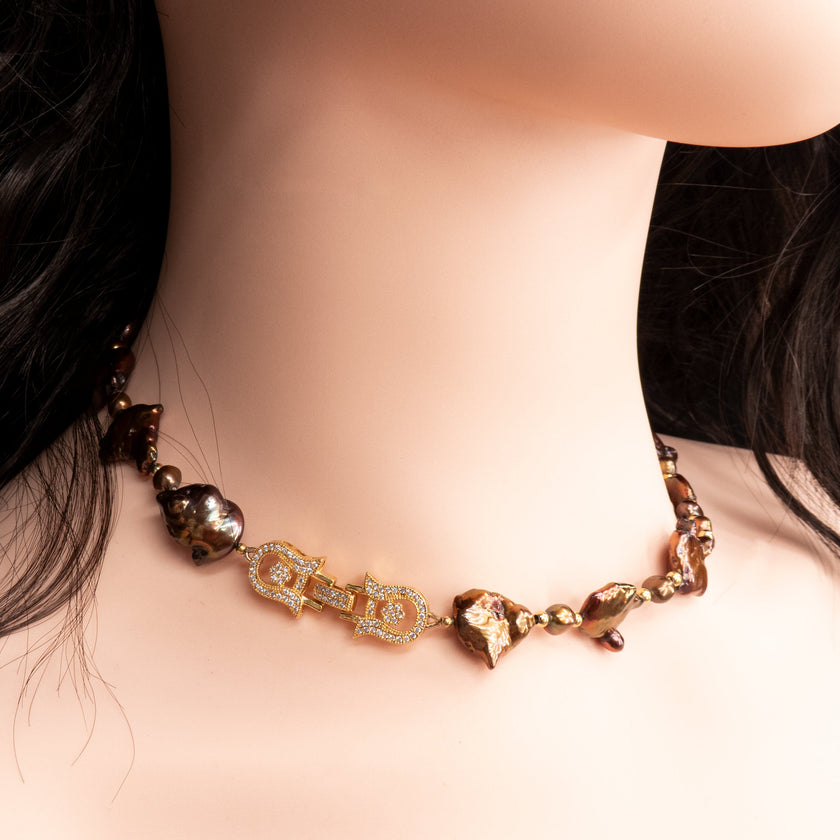 שרשרת נדירה שמדברת בעד עצמה המורכבת מפניני בארוק טבעיות בגוון שוקולד עמוק, עם אביזרים בציפוי זהב שמדגישים את צורתן הבלתי אחידה והמרקם החי של כל פנינה. הברק המטאלי החם של הפנינים משתלב באופן הרמוני עם הנגיעות המוזהבות, ויוצר מראה חם ועשיר שמזכיר אדמה וקליפה. שילוב קלאסי ונועז כאחד – אידיאלי לאירוע ערב, להופעה עם אמירה או כמתנה מרשימה לאישה מיוחדת. פניני הבארוק, כל אחת עם אישיות משלה, הופכות את השרשרת הזו לתכשיט שמבליט את הייחוד שלך בלי מילים.