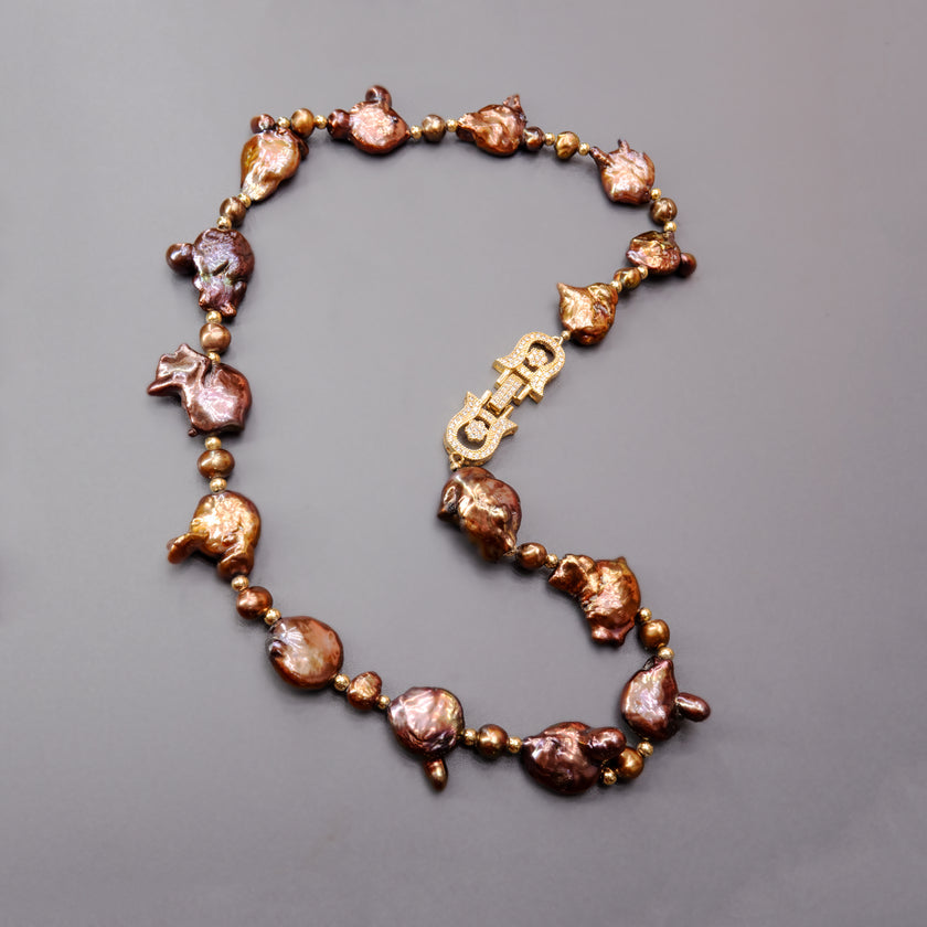 תכשיט בולט עם אופי טבעי המורכבת מפניני בארוק טבעיות בגוון שוקולד עמוק, עם אביזרים בציפוי זהב שמדגישים את צורתן הבלתי אחידה והמרקם החי של כל פנינה. הברק המטאלי החם של הפנינים משתלב באופן הרמוני עם הנגיעות המוזהבות, ויוצר ניחוח של טבע פראי ואלגנטי יחד. שילוב בין חוזק טבעי ליוקרה נינוחה – אידיאלי לאירוע ערב, להופעה עם אמירה או כמתנה מרשימה לאישה מיוחדת. פניני הבארוק, כל אחת עם אישיות משלה, הופכות את השרשרת הזו לשרשרת שלא דומה לשום דבר אחר – טבעית, אותנטית ומרשימה.