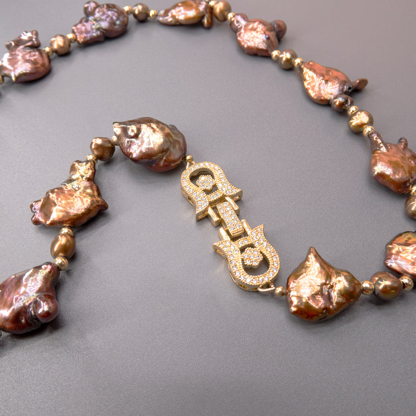 תכשיט בולט עם אופי טבעי המורכבת מפניני בארוק טבעיות בגוון שוקולד עמוק, עם אביזרים בציפוי זהב שמדגישים את צורתן הבלתי אחידה והמרקם החי של כל פנינה. הברק המטאלי החם של הפנינים משתלב באופן הרמוני עם הנגיעות המוזהבות, ויוצר מראה חם ועשיר שמזכיר אדמה וקליפה. שילוב קלאסי ונועז כאחד – אידיאלי לאירוע ערב, להופעה עם אמירה או כמתנה מרשימה לאישה מיוחדת. פניני הבארוק, כל אחת עם אישיות משלה, הופכות את השרשרת הזו לשרשרת שלא דומה לשום דבר אחר – טבעית, אותנטית ומרשימה.