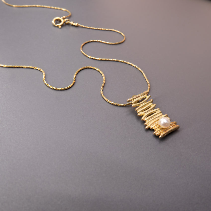 צילום תקריב של שרשרת עם תליון בעיצוב אורגני מופשט מזהב, המדמה שכבות טבעיות עם נגיעת טקסטורה. במרכז – פנינה לבנה שמוסיפה ברק ועדינות. שילוב מרהיב של ניגודים: מתכת ופנינה, גולמי ורך, מודרני ועל זמני.