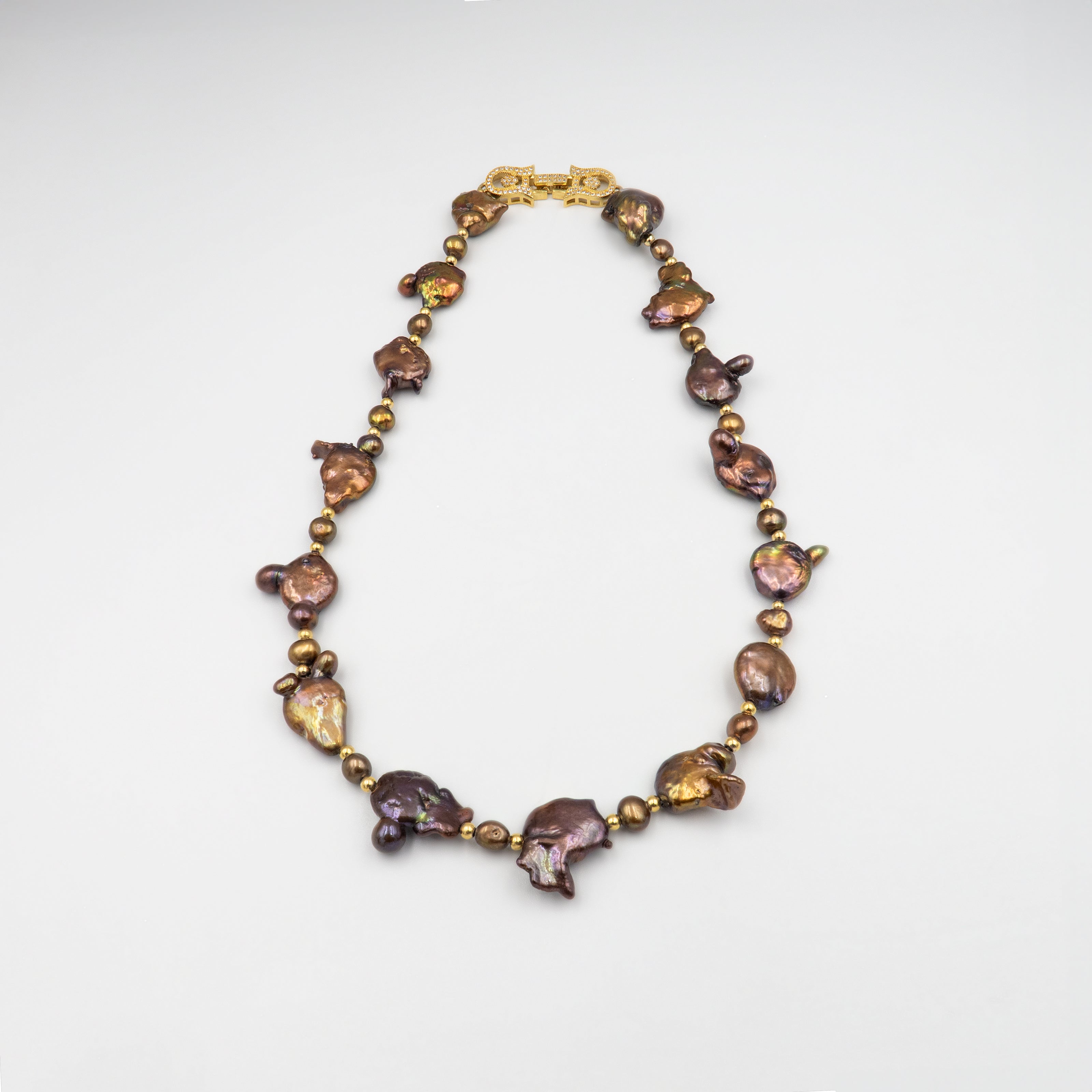 פריט בעיצוב לא שגרתי, מלא נוכחות המורכבת מפניני בארוק טבעיות בגוון שוקולד עמוק, עם אביזרים בציפוי זהב שמדגישים את צורתן הבלתי אחידה והמרקם החי של כל פנינה. הברק המטאלי החם של הפנינים משתלב באופן הרמוני עם הנגיעות המוזהבות, ויוצר תחושה של חומר חי ונושם. השראה מהטבע עם קריצה עיצובית מודרנית – אידיאלי לאירוע ערב, להופעה עם אמירה או כמתנה מרשימה לאישה מיוחדת. פניני הבארוק, כל אחת עם אישיות משלה, הופכות את השרשרת הזו לתכשיט שמבליט את הייחוד שלך בלי מילים.