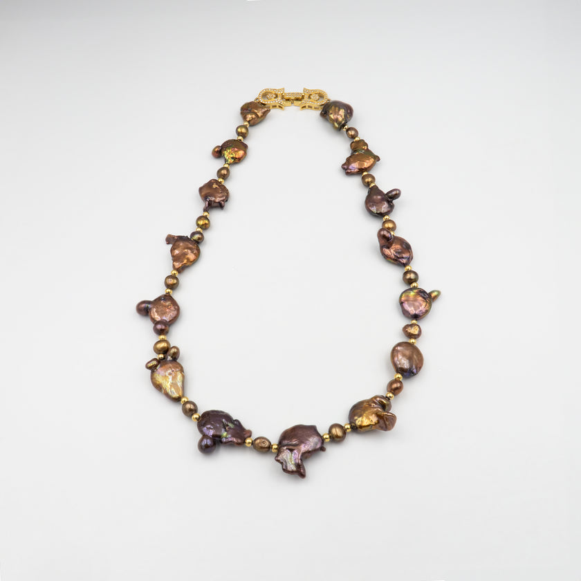 פריט בעיצוב לא שגרתי, מלא נוכחות המורכבת מפניני בארוק טבעיות בגוון שוקולד עמוק, עם אביזרים בציפוי זהב שמדגישים את צורתן הבלתי אחידה והמרקם החי של כל פנינה. הברק המטאלי החם של הפנינים משתלב באופן הרמוני עם הנגיעות המוזהבות, ויוצר תחושה של חומר חי ונושם. השראה מהטבע עם קריצה עיצובית מודרנית – אידיאלי לאירוע ערב, להופעה עם אמירה או כמתנה מרשימה לאישה מיוחדת. פניני הבארוק, כל אחת עם אישיות משלה, הופכות את השרשרת הזו לתכשיט שמבליט את הייחוד שלך בלי מילים.