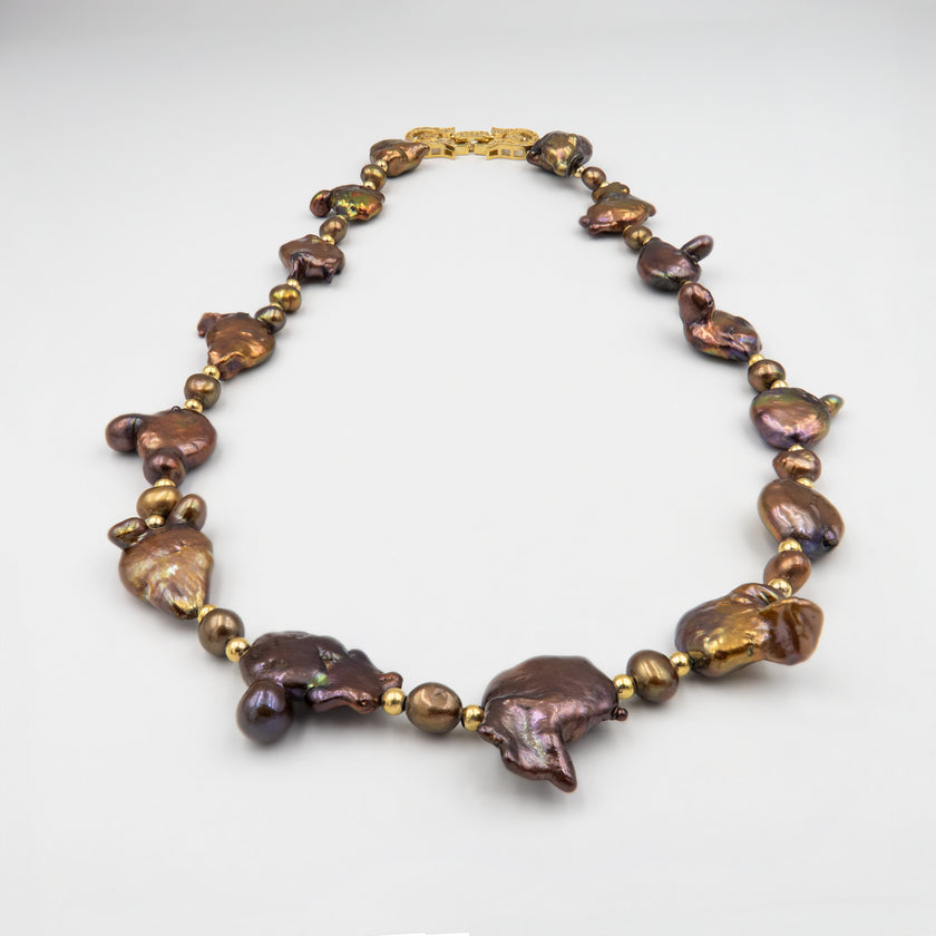 שרשרת נדירה שמדברת בעד עצמה המורכבת מפניני בארוק טבעיות בגוון שוקולד עמוק, עם אביזרים בציפוי זהב שמדגישים את צורתן הבלתי אחידה והמרקם החי של כל פנינה. הברק המטאלי החם של הפנינים משתלב באופן הרמוני עם הנגיעות המוזהבות, ויוצר ניחוח של טבע פראי ואלגנטי יחד. שילוב קלאסי ונועז כאחד – אידיאלי לאירוע ערב, להופעה עם אמירה או כמתנה מרשימה לאישה מיוחדת. פניני הבארוק, כל אחת עם אישיות משלה, הופכות את השרשרת הזו לשרשרת שלא דומה לשום דבר אחר – טבעית, אותנטית ומרשימה.