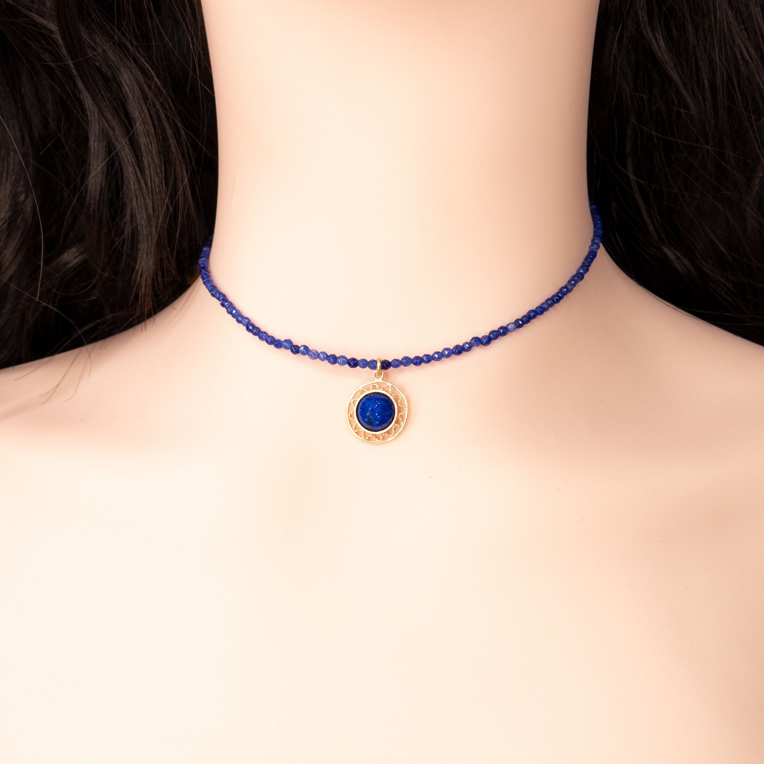 שרשרת קולר עדינה עשויה חרוזים קטנים בגוון כחול רויאל עמוק, עם תליון עגול מוזהב במרכזו, שבתוכו שיבוץ אבן כחולה מרהיבה. התליון מעוטר במסגרת חרוטה דקיקה וזהובה, ויושב בצורה מדויקת בקו הצוואר. התכשיט מצולם על צוואר של בובת תצוגה, ברקע נייטרלי המדגיש את צבעי התכשיט. שילוב של קלאסיות עוצמתית עם עיצוב מינימליסטי.