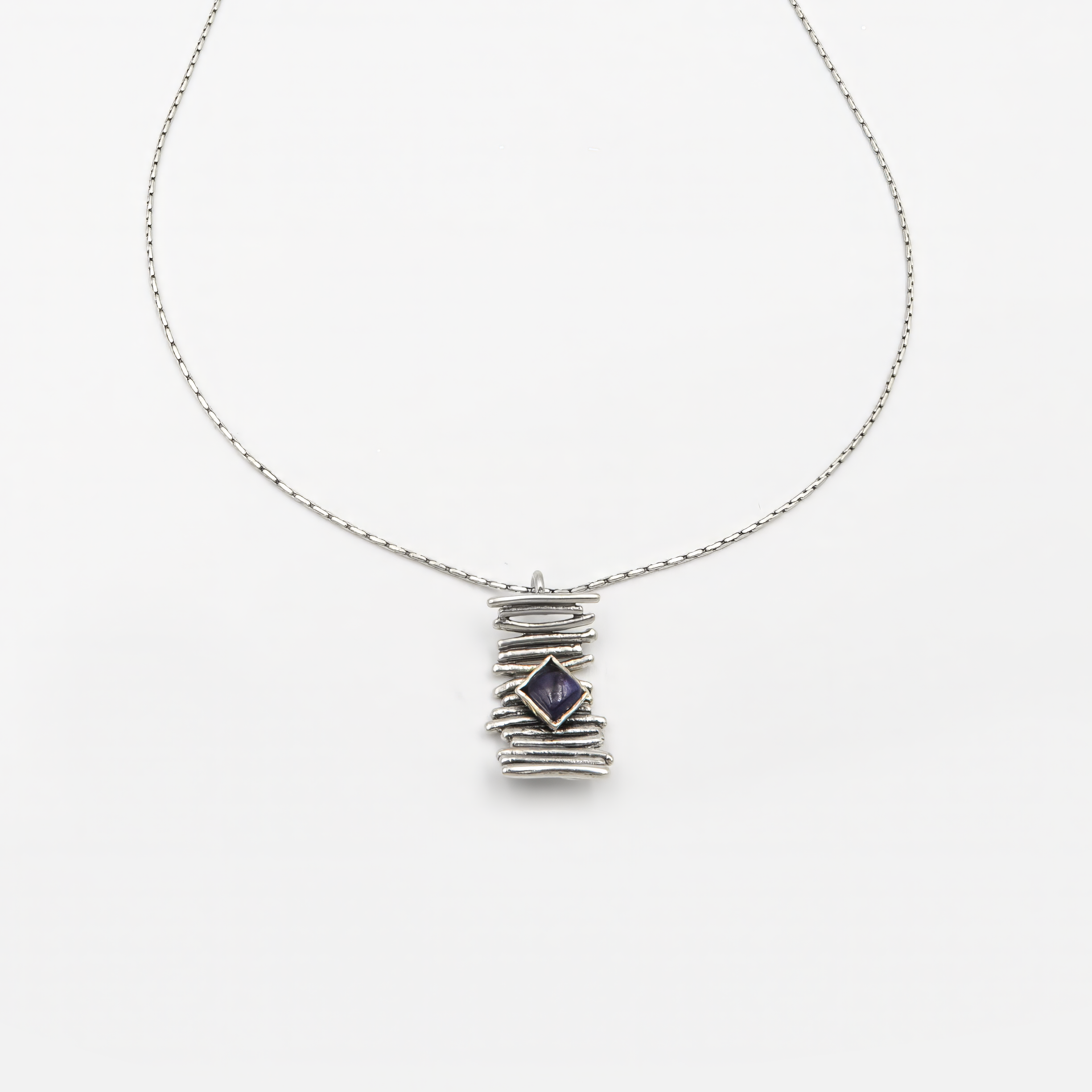 שרשרת כסף עדינה עם שורת חרוזים כסופים מבריקים במרכז, שמתחתיהם תלוי תליון קטן ועגול המשובץ באבן איולייט טבעית. האבן מציגה גוון כחול-סגלגל עמוק עם ברק פנימי שקוף, אופייני לאיולייט – אבן הידועה בזוהר המשתנה שלה בהתאם לזווית התאורה. מסגרת התליון חלקה ועגולה, ומעניקה לאבן במה נקייה ונוכחת. עיצוב שמאזן בין פשטות גיאומטרית ליופי טבעי ומסתורי.