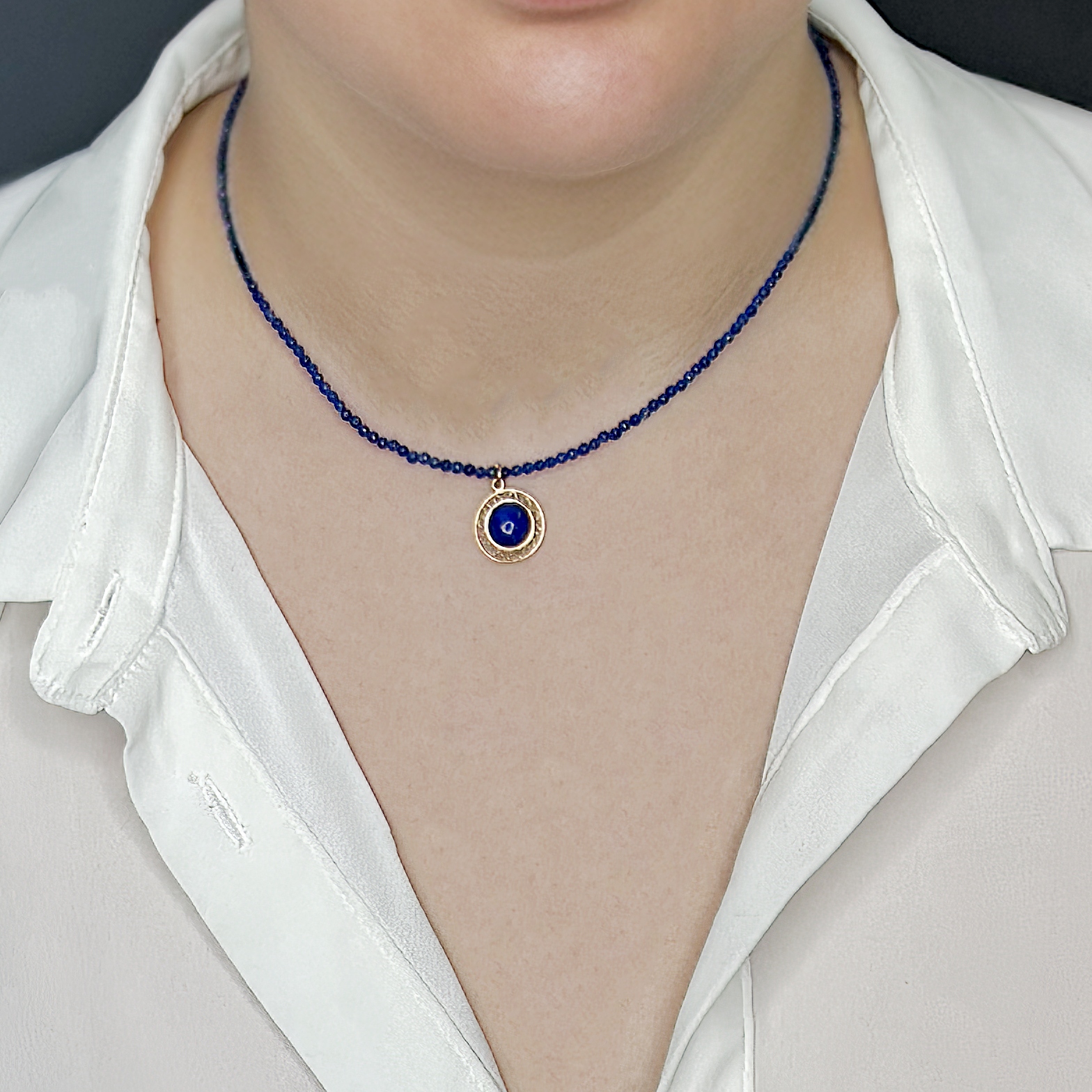 שרשרת צ’וקר קצרה עשויה חרוזים קטנים ואחידים בגוון כחול כהה, עם תליון מוזהב עגול במרכזו שיבוץ אבן כחולה דמוית לאפיס לאזולי. מסגרת התליון חרוטה בעיטור גאומטרי עדין. התכשיט מונח בצורה סימטרית סביב הצוואר ונראה מעוצב בקפידה. תכשיט בעיצוב מינימליסטי אך בעל נוכחות, מושלם לשילוב עם לוקים מודרניים.

