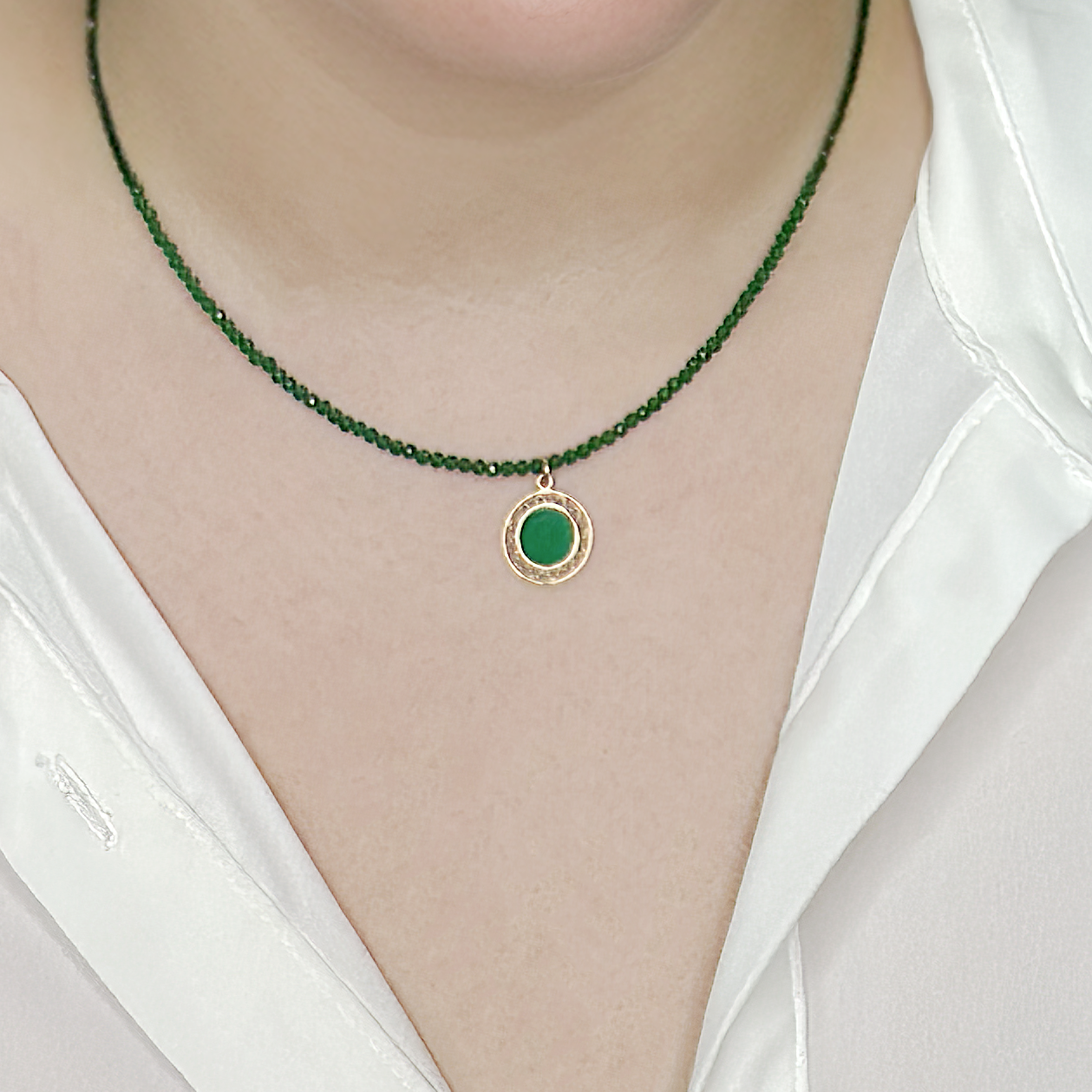 שרשרת קולר אלגנטית עשויה חרוזים ירוקים קטנים ומבריקים, עם תליון עגול מוזהב במרכזו, שבתוכו משובצת אבן ירוקה בעלת ברק עמום. השרשרת מונחת בעדינות על הצוואר ויוצרת ניגוד מרשים מול חולצה לבנה חלקה. עיצוב קלאסי שמתאים ליום ולערב, עם מראה נקי ויוקרתי.