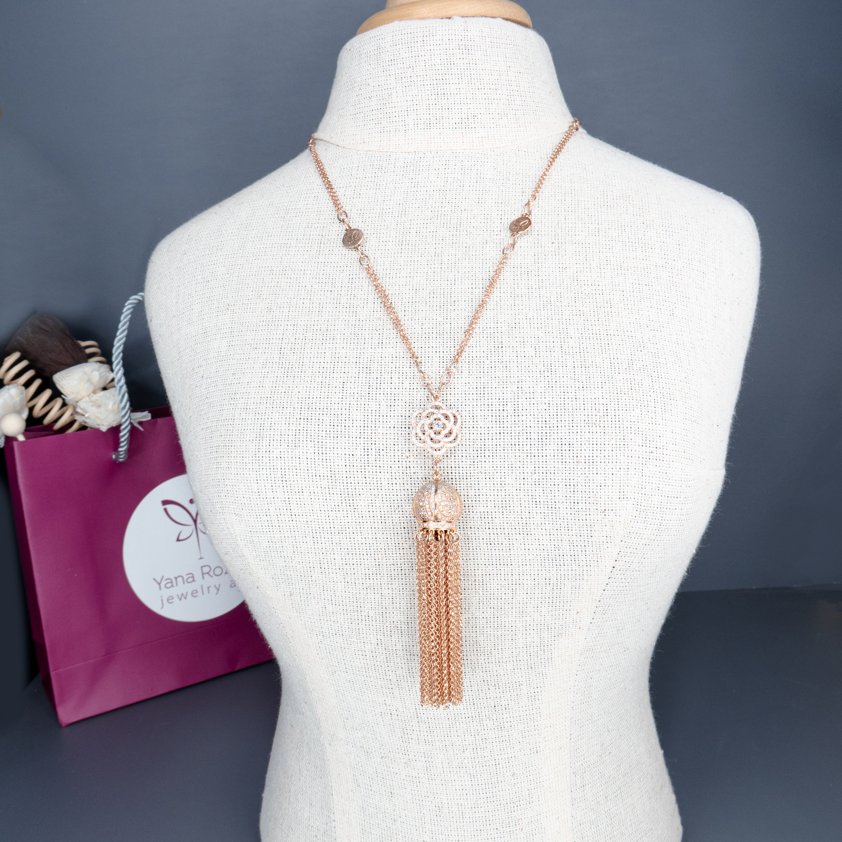 שרשרת ארוכה בציפוי זהב ורוד עם תליון מרכזי מעוטר בעיצוב פרחוני עדין ודמוי ורד, ומשתרכת ממנו כדור תחרה מתכתי שממנו משתלשלות שורות של שרשראות דקיקות. השרשרת מוצגת על בובת תצוגה בגוון בז', ברקע שקית סגולה עם לוגו 