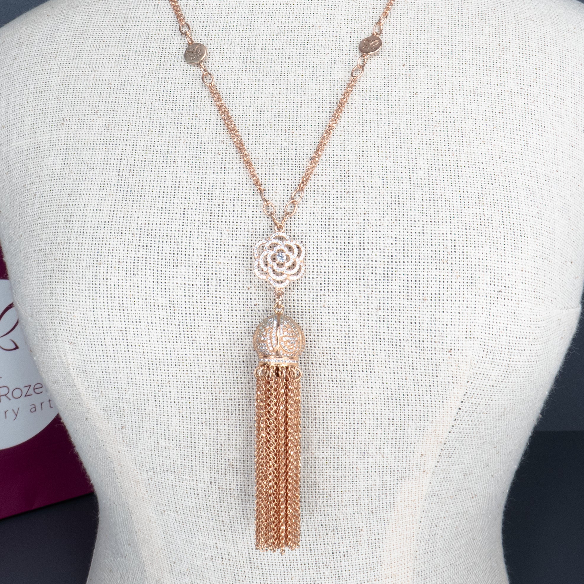 שרשרת אלגנטית בגוון רוז גולד עם אלמנט מרכזי בצורת פרח תחרה מתכתי, וכדור משובץ ממנו משתלשלת פיסת גדילים ארוכה משרשראות דקות. מוצג על דגם תצוגה עם טקסטורת פשתן. מאחור נראית שקית ממותגת של 