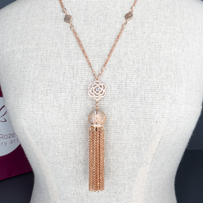 שרשרת אלגנטית בגוון רוז גולד עם אלמנט מרכזי בצורת פרח תחרה מתכתי, וכדור משובץ ממנו משתלשלת פיסת גדילים ארוכה משרשראות דקות. מוצג על דגם תצוגה עם טקסטורת פשתן. מאחור נראית שקית ממותגת של 