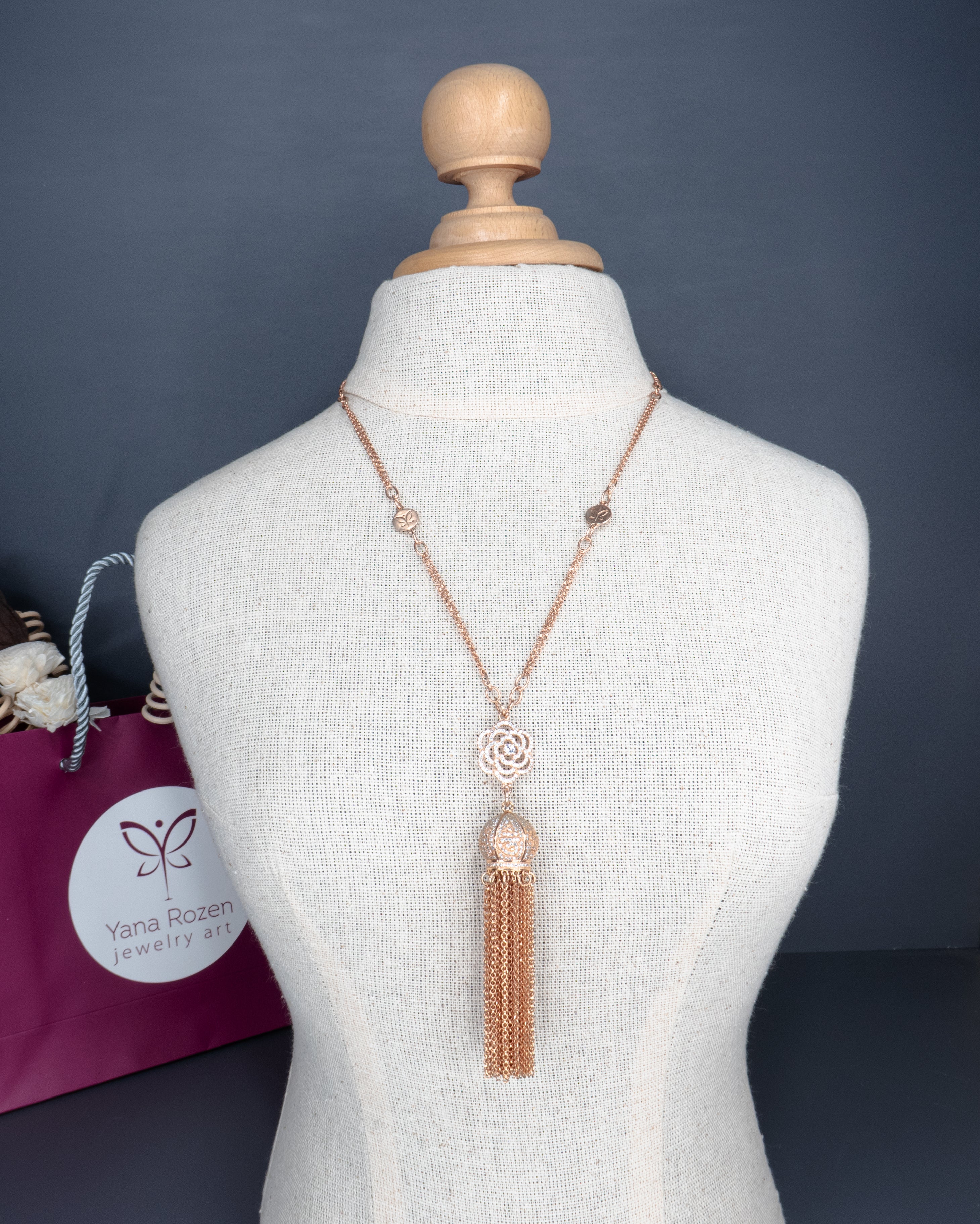 תכשיט רומנטי ומוקפד: שרשרת ארוכה בגוון זהב ורוד עם עיצוב מרכזי של פרח פתוח, כדור דקורטיבי וגדילים רבים שמשתלשלים כלפי מטה. מוצגת על בובת תצוגה אלגנטית, עם שקית תכשיטים יוקרתית בצבע בורדו כהה ברקע. עיצוב שמשדר נשיות קלאסית עם נגיעות מודרניות.