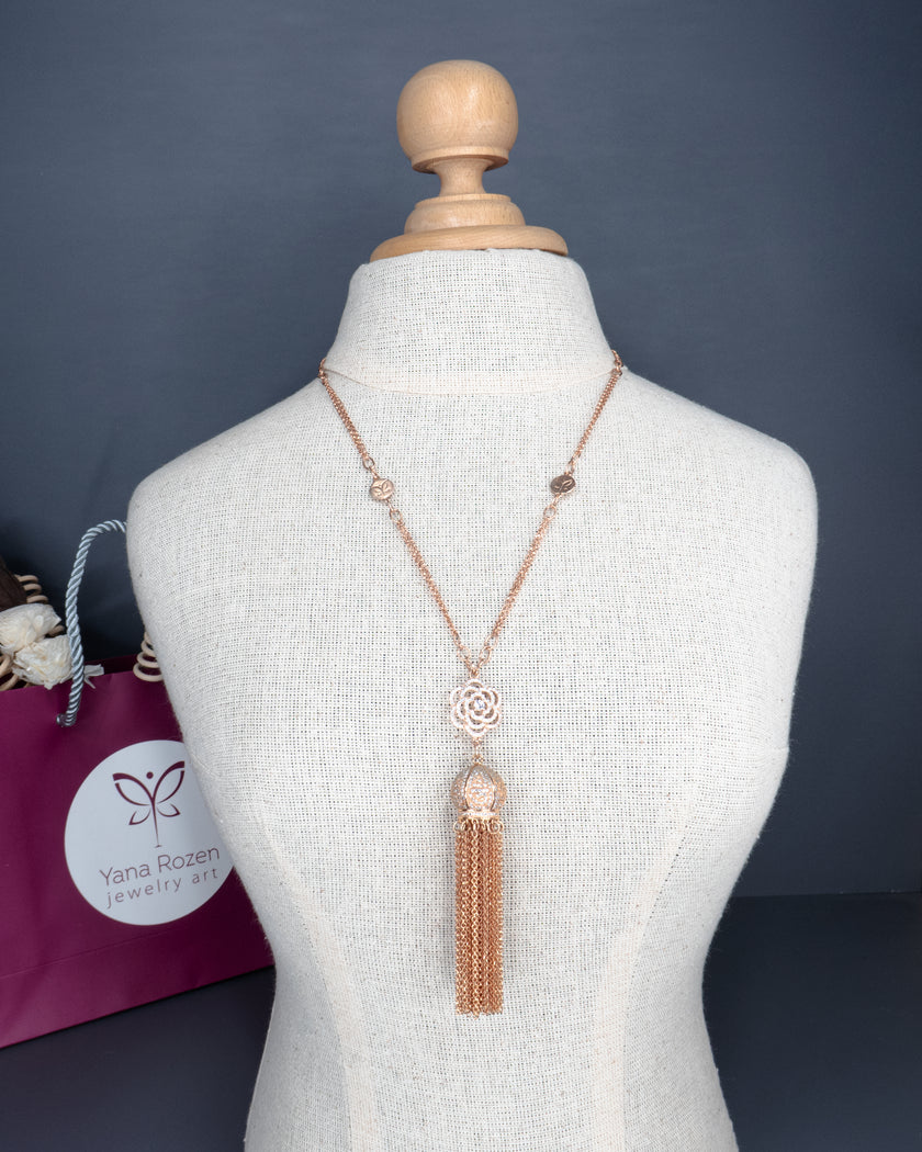 תכשיט רומנטי ומוקפד: שרשרת ארוכה בגוון זהב ורוד עם עיצוב מרכזי של פרח פתוח, כדור דקורטיבי וגדילים רבים שמשתלשלים כלפי מטה. מוצגת על בובת תצוגה אלגנטית, עם שקית תכשיטים יוקרתית בצבע בורדו כהה ברקע. עיצוב שמשדר נשיות קלאסית עם נגיעות מודרניות.