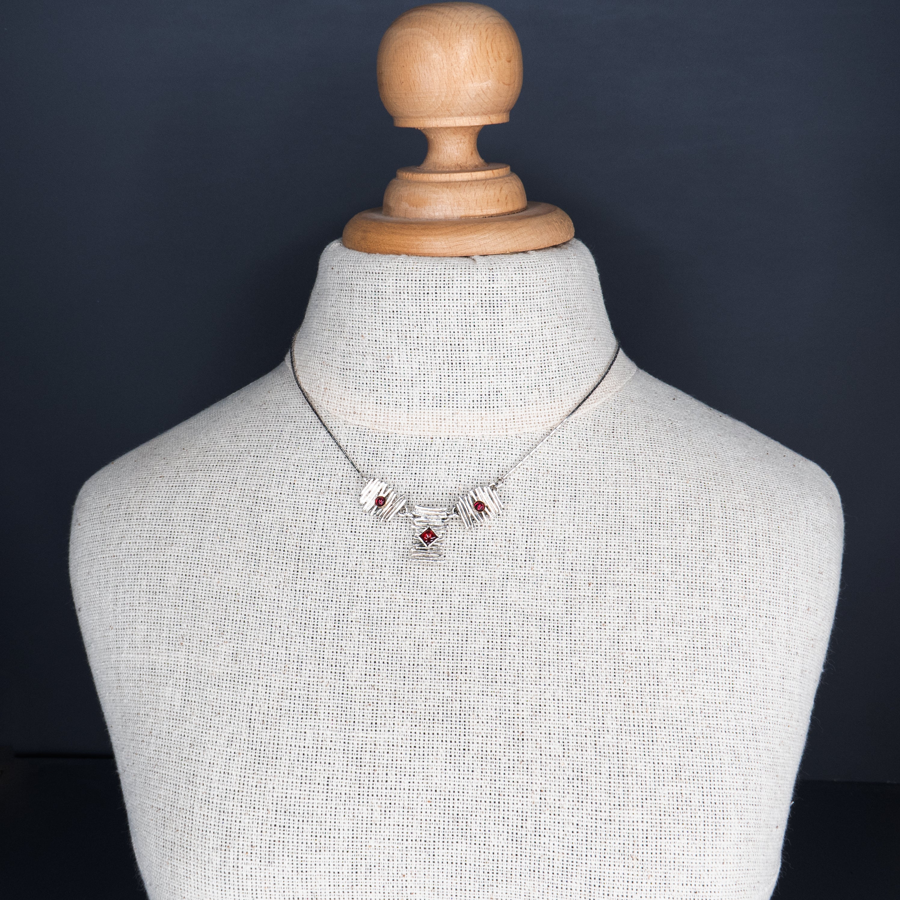 שרשרת ברוטליסטית בהשראת שנות ה-70: לוחות כסף סטרלינג דמויי קיסמים מוערמים בשלושה מקטעים. בלוח המרכזי שיבוץ גרנט חיתוך נסיכה, ובשניים הצדדיים גרנטים עגולים קטנים. הלוחות מחוברים לשרשרת דקיקה, והניגוד בין המרקם המחוספס לכוחו של האדום העמוק יוצר חזות דרמטית אך אלגנטית.