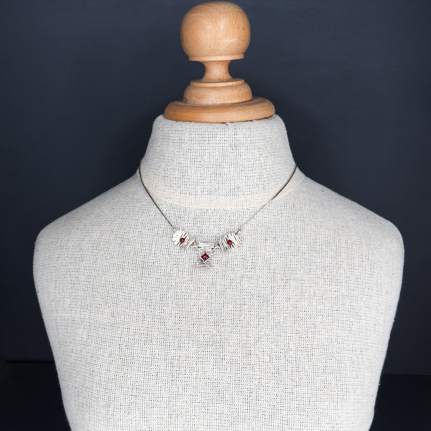 שרשרת ברוטליסטית בהשראת שנות ה-70: לוחות כסף סטרלינג דמויי קיסמים מוערמים בשלושה מקטעים. בלוח המרכזי שיבוץ גרנט חיתוך נסיכה, ובשניים הצדדיים גרנטים עגולים קטנים. הלוחות מחוברים לשרשרת דקיקה, והניגוד בין המרקם המחוספס לכוחו של האדום העמוק יוצר חזות דרמטית אך אלגנטית.